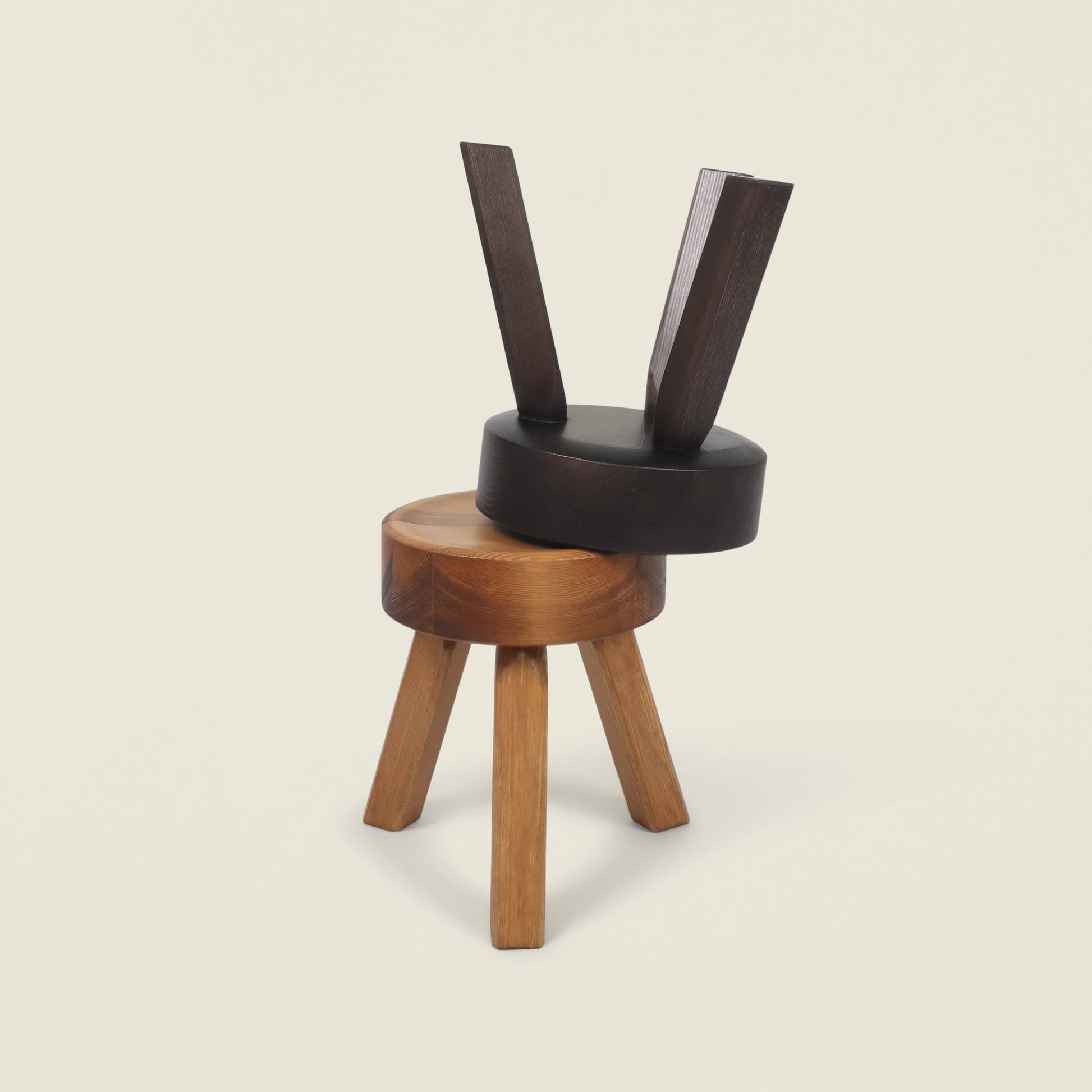 Two FRAMA AML Stools from FRAMA—one solid oiled pine three-legged stool upright, the other dark wood at an angle—are stacked in a sculptural arrangement, highlighting their versatile design against a plain background.