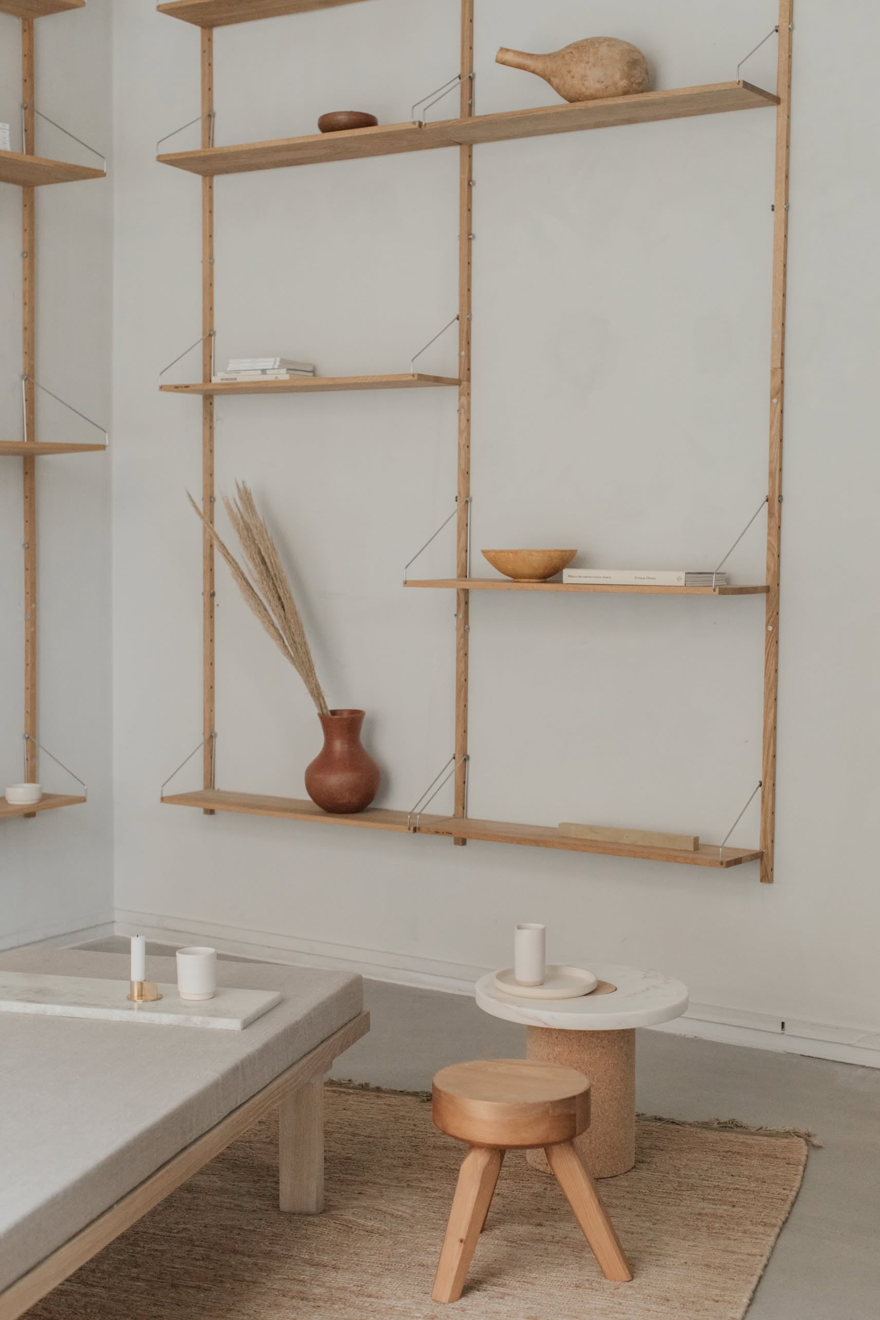 A minimalist living space features wooden wall shelves with pottery, a brown vase, bowls, books, and candles. The versatile FRAMA AML Stool by FRAMA serves as a bedside table alongside a round table and a light floor rug.
