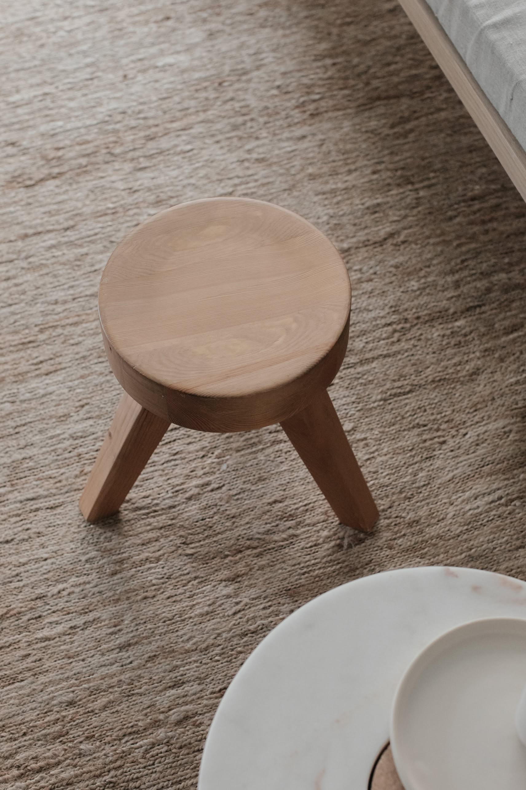 The FRAMA AML Stool by FRAMA, crafted from solid oiled pine with a versatile design, sits on a textured beige carpet beside a white round table and near a bed.