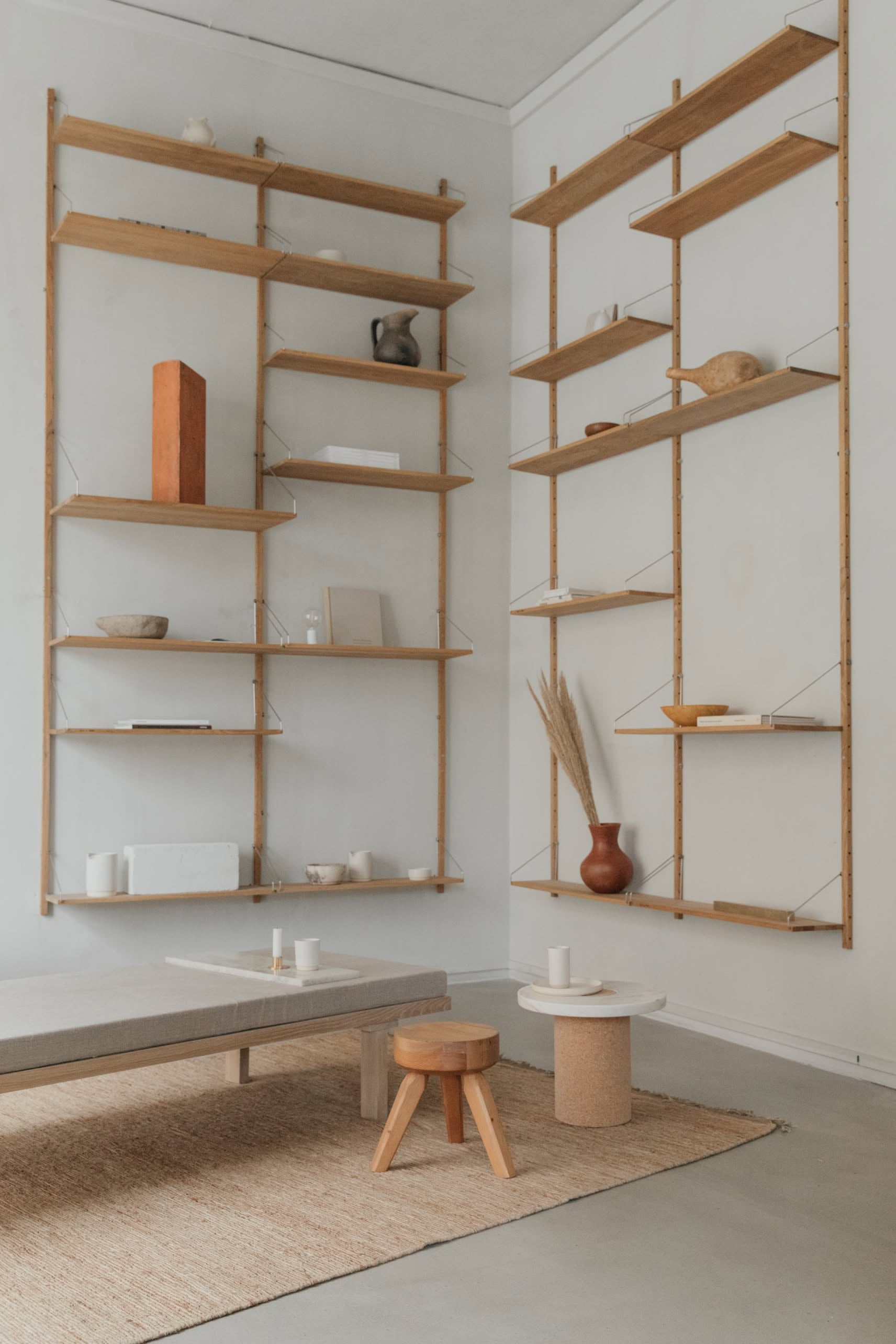 Minimalist living room with light wood wall shelves, a low cushioned bench, and a round side table holding a vase of dried grass. The versatile FRAMA AML Stool stands out beside a jute rug on the concrete floor.