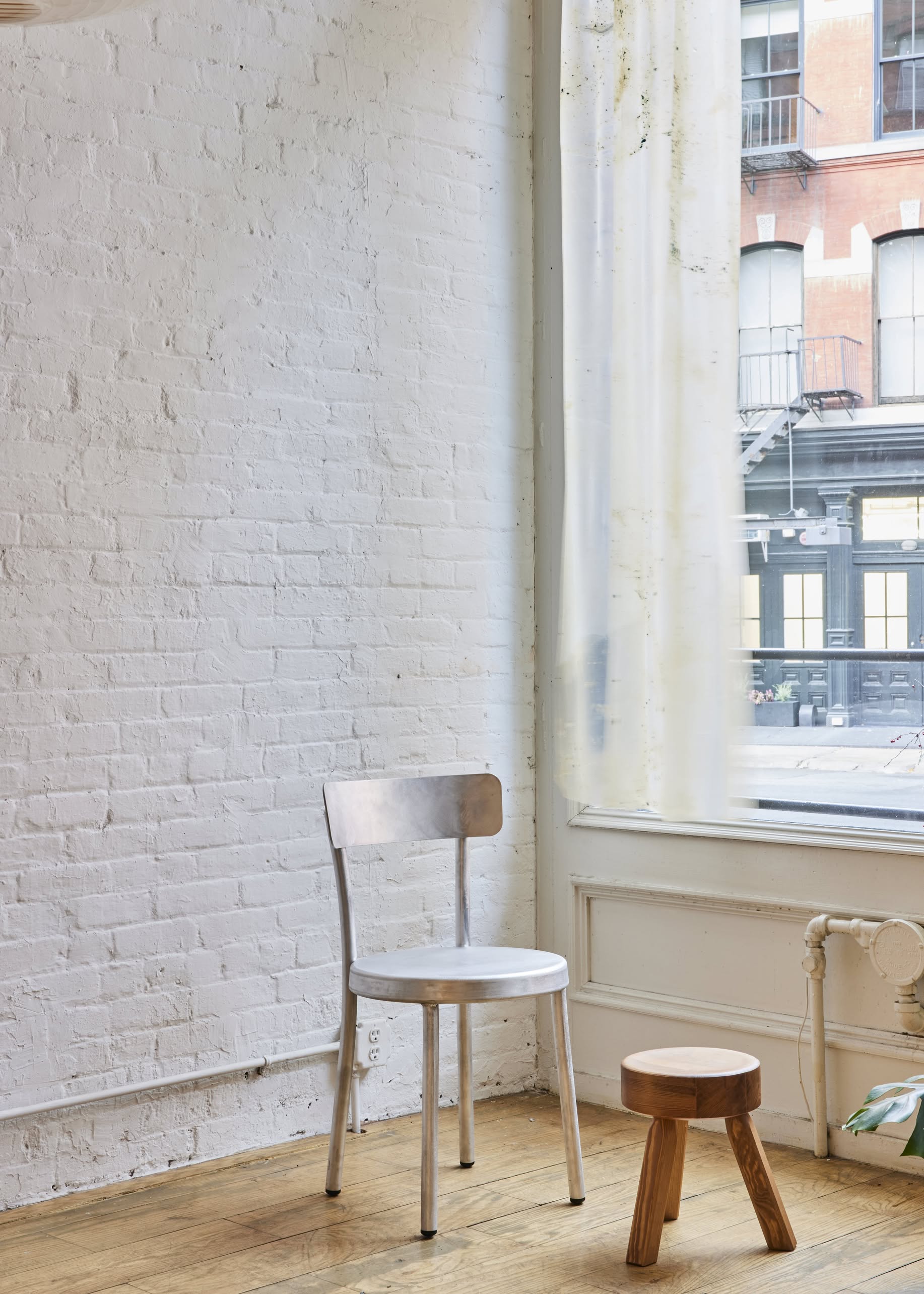 A FRAMA AML Stool by FRAMA stands on a light wood floor near a large window with sheer curtains in a bright room with white brick walls, overlooking a city street.