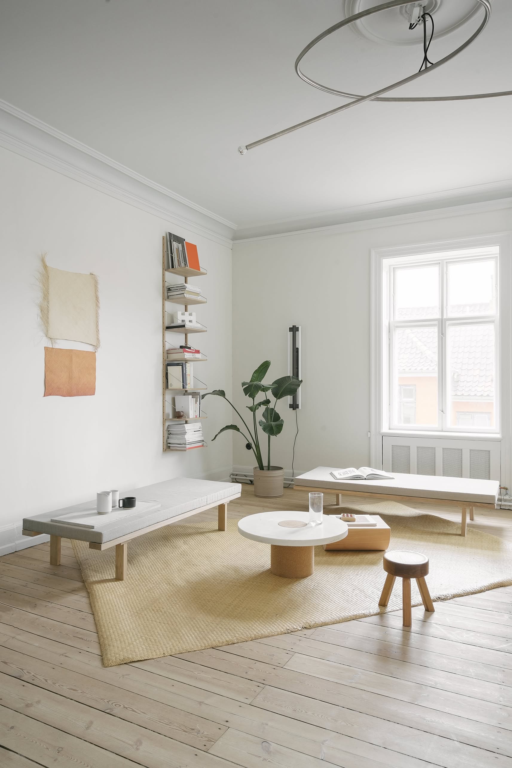 Minimalist living room with light wood floors, low modern furniture, a round coffee table, the FRAMA AML Stool in solid oiled pine, a potted plant, wall shelves with books, versatile decor, large window, and soft natural lighting.