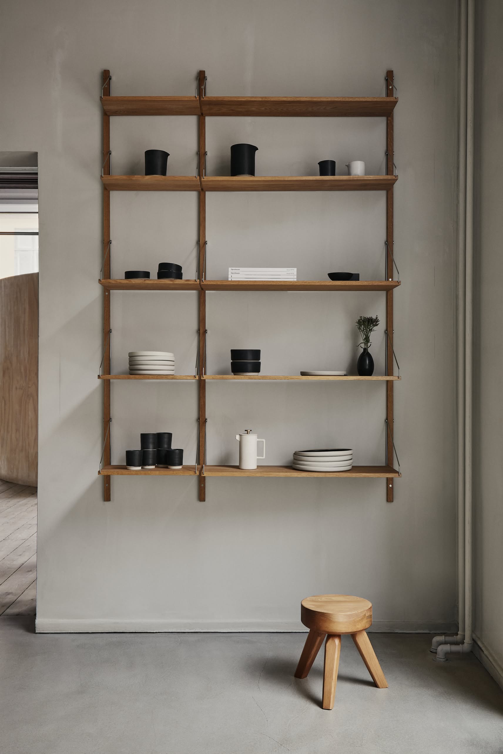 A solid oiled pine wall-mounted shelving unit displays black and white dishes, cups, bowls, and a small potted plant above a FRAMA AML Stool by FRAMA, set on a concrete floor against a gray wall.