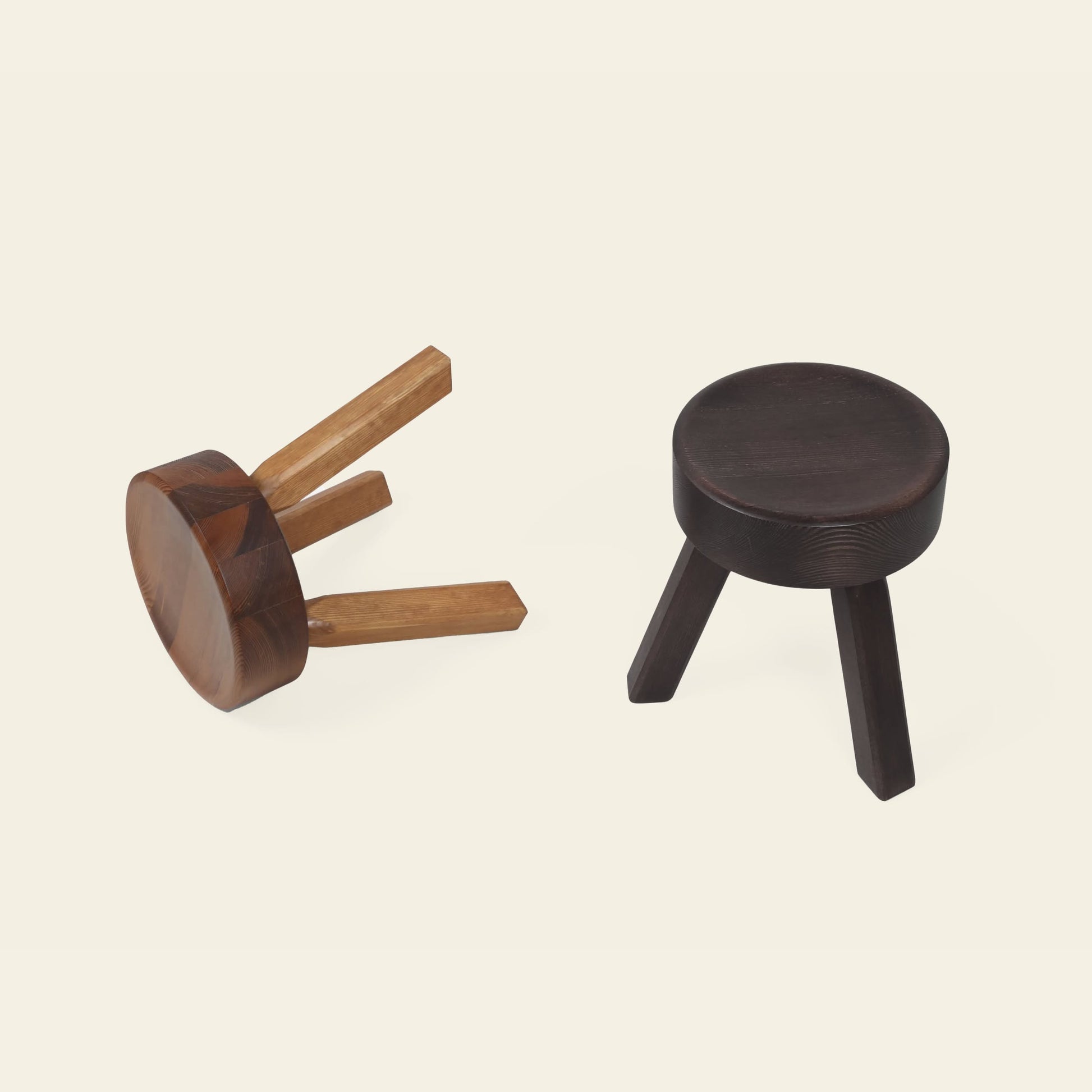 Two three-legged FRAMA AML Stools by FRAMA, crafted from solid oiled pine—one upright in dark brown, the other lighter brown and lying on its side—highlight their versatile design against a plain off-white background.