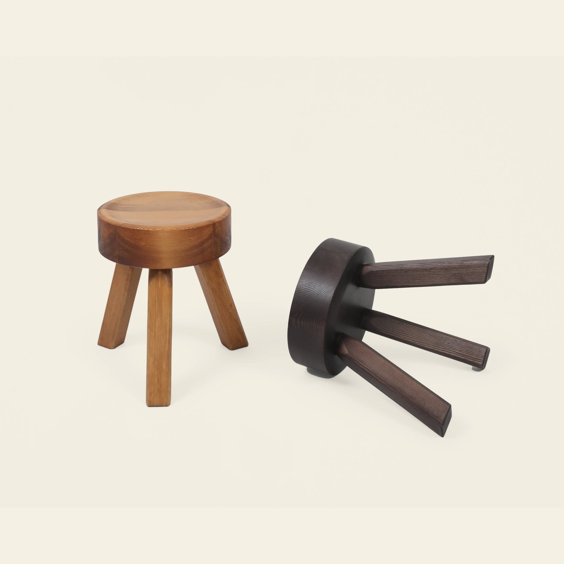 Two FRAMA AML Stools by FRAMA, each with three legs and crafted from solid oiled pine, are displayed—one upright in light wood and the other dark and on its side—showcasing their versatile use as bedside stools or seating.