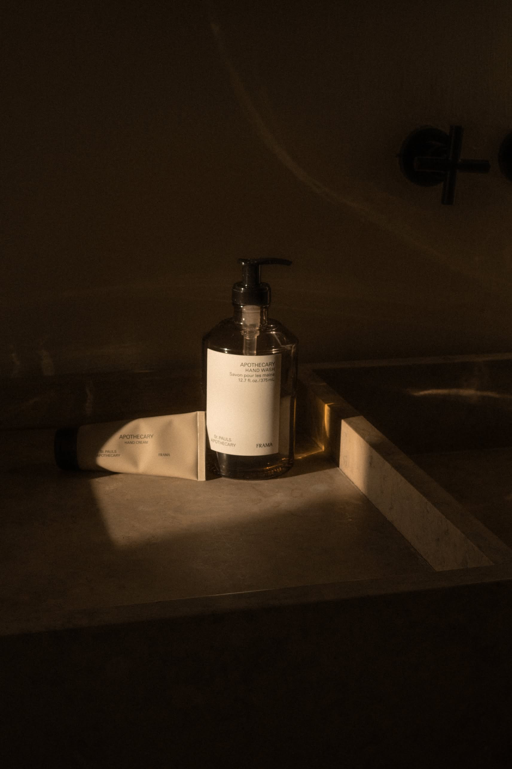 A 375ml FRAMA Hand Wash pump bottle and a skincare tube rest on a marble sink, bathed in warm, soft light in a dimly lit bathroom.