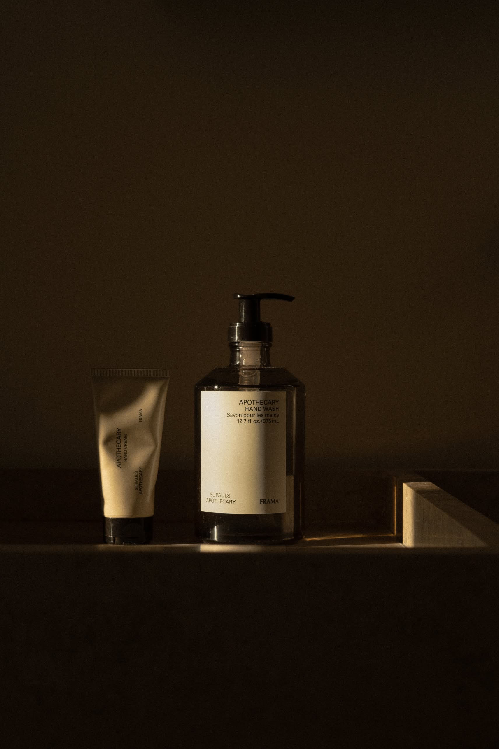 Two FRAMA skincare products—a tube and a 375ml Hand Wash pump—rest side by side in dim, moody lighting. The partially visible labels suggest organic ingredients and sandalwood scent but aren’t fully readable.