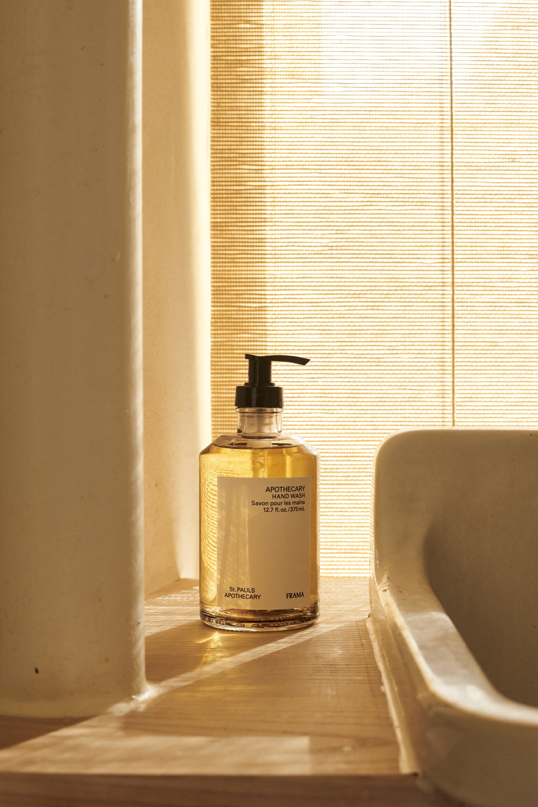A 375ml FRAMA Hand Wash with a pump rests by the sink in warm sunlight, near a bamboo-shaded window and beige wall, releasing a soothing sandalwood scent from its organic ingredients.