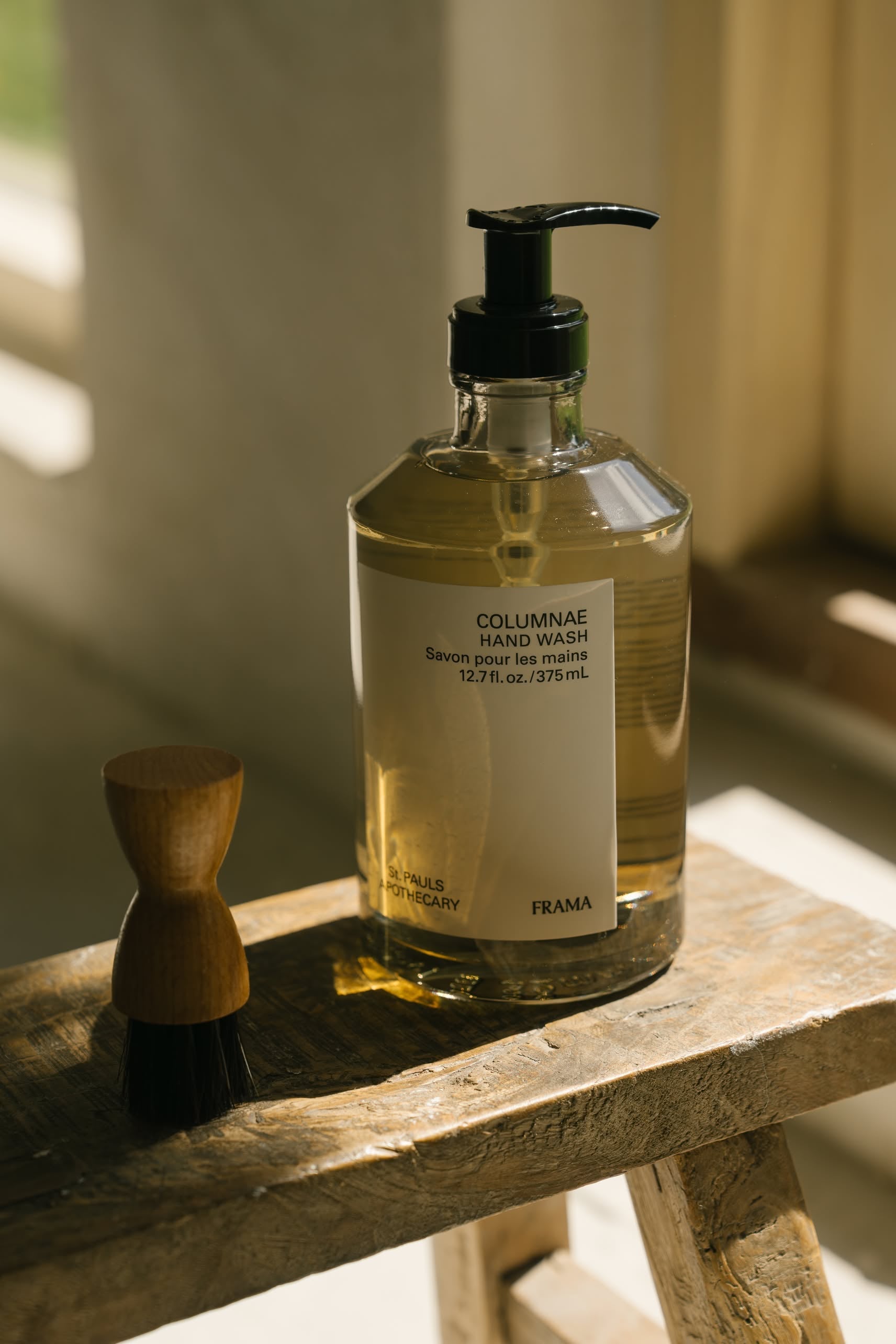 A clear 375ml bottle of FRAMA Hand Wash with a black pump sits on a rustic wooden surface beside a small wooden-handled brush, illuminated by natural sunlight.