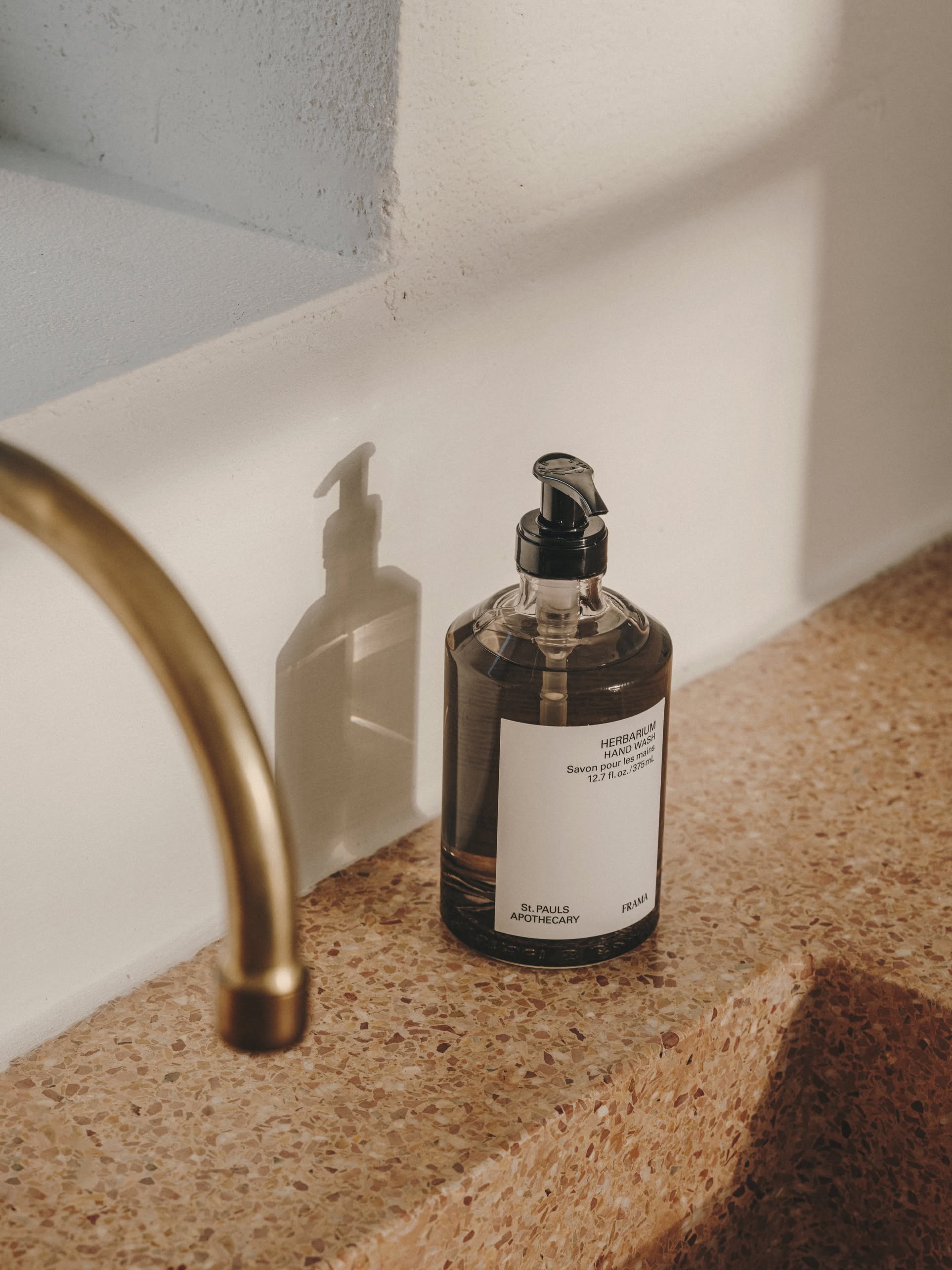 A 375ml FRAMA Hand Wash bottle with pump dispenser rests on a terrazzo countertop beside a brass faucet, casting a shadow on a white wall in gentle natural light.