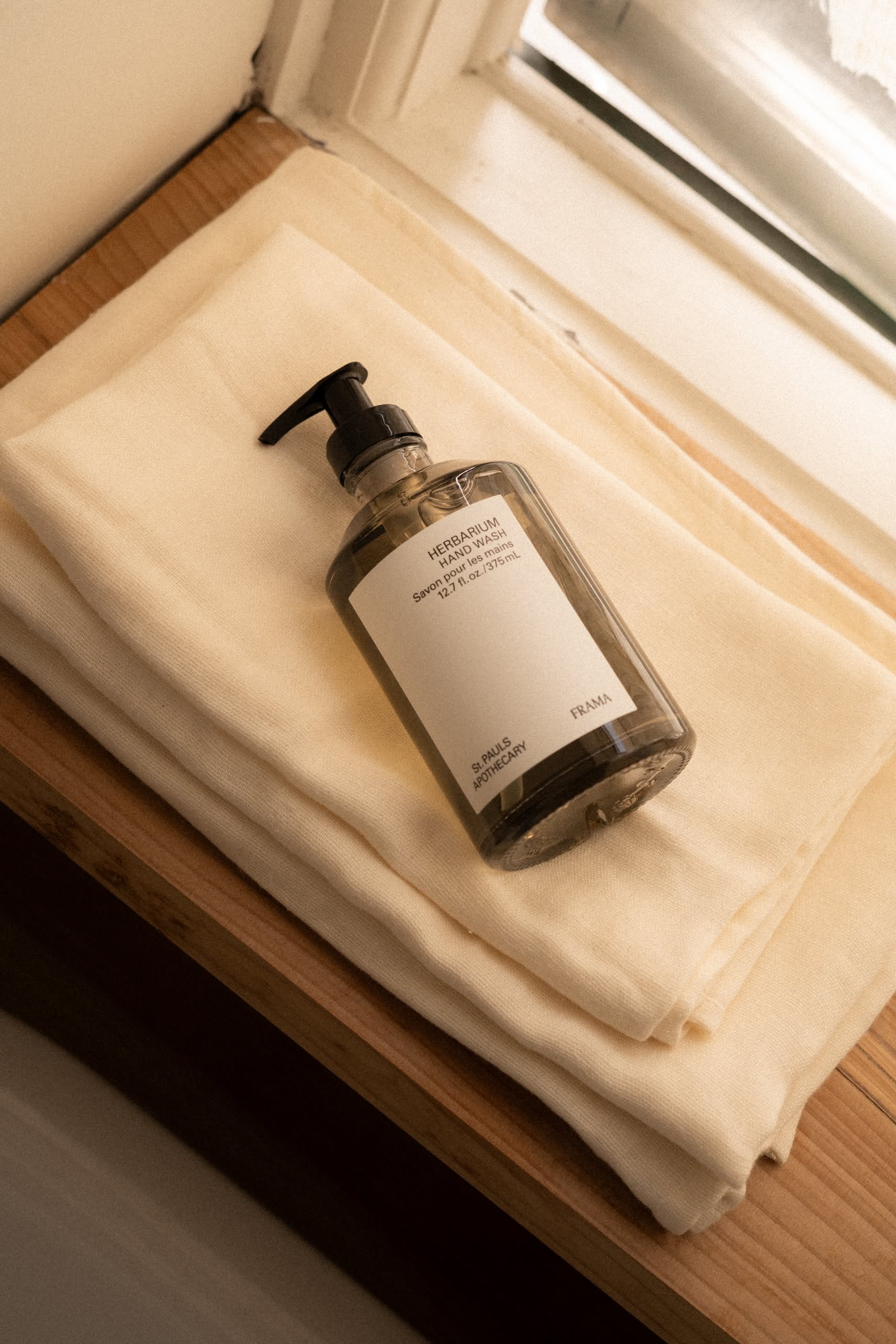 A 375ml brown pump bottle of FRAMA Hand Wash with a white label sits on folded cream towels atop a wooden surface near a window, bathed in natural light.