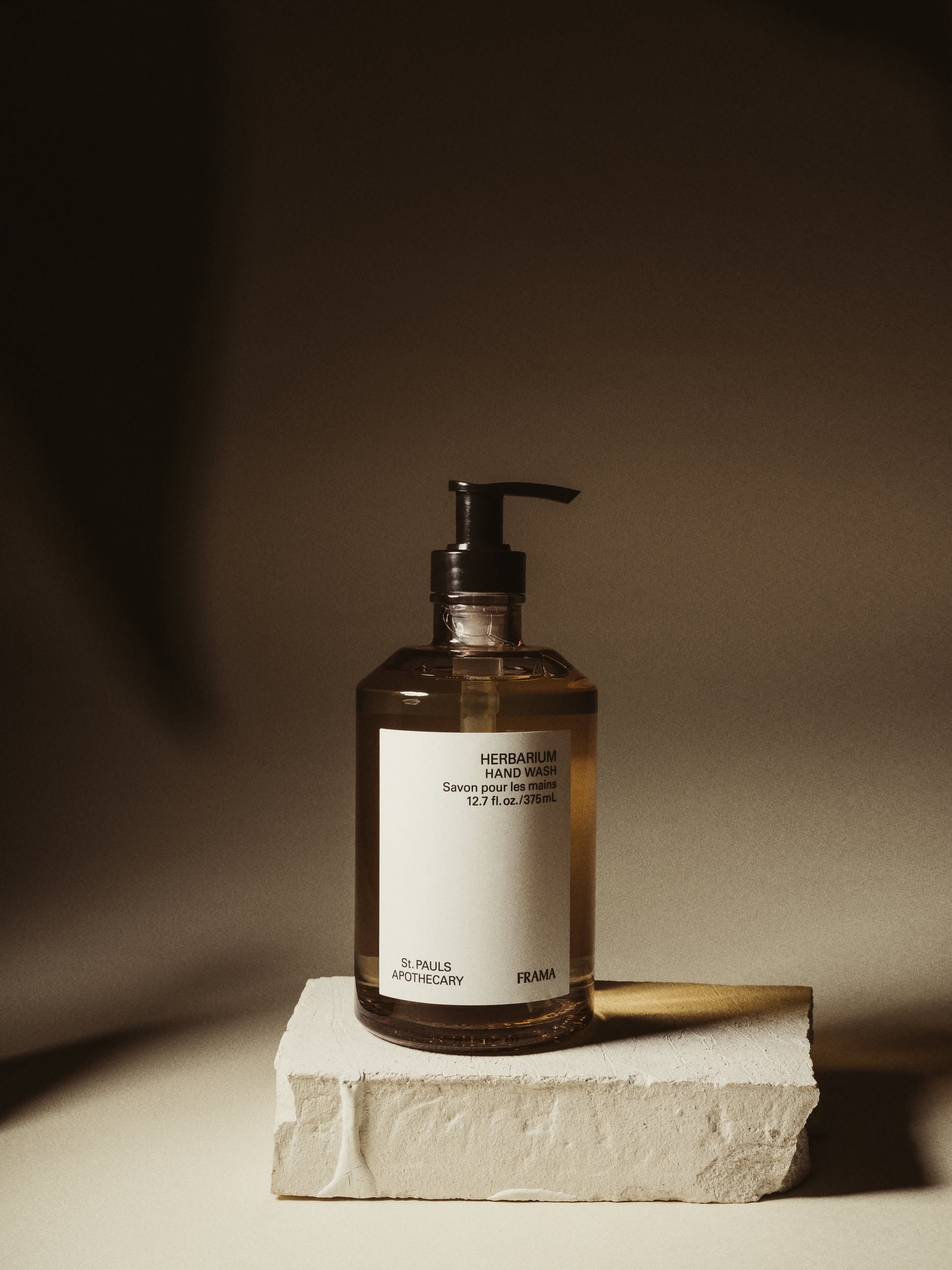 A clear pump bottle of FRAMA Hand Wash, 375ml, featuring the FRAMA label and organic ingredients, is placed on a textured white block against a softly lit brown background.