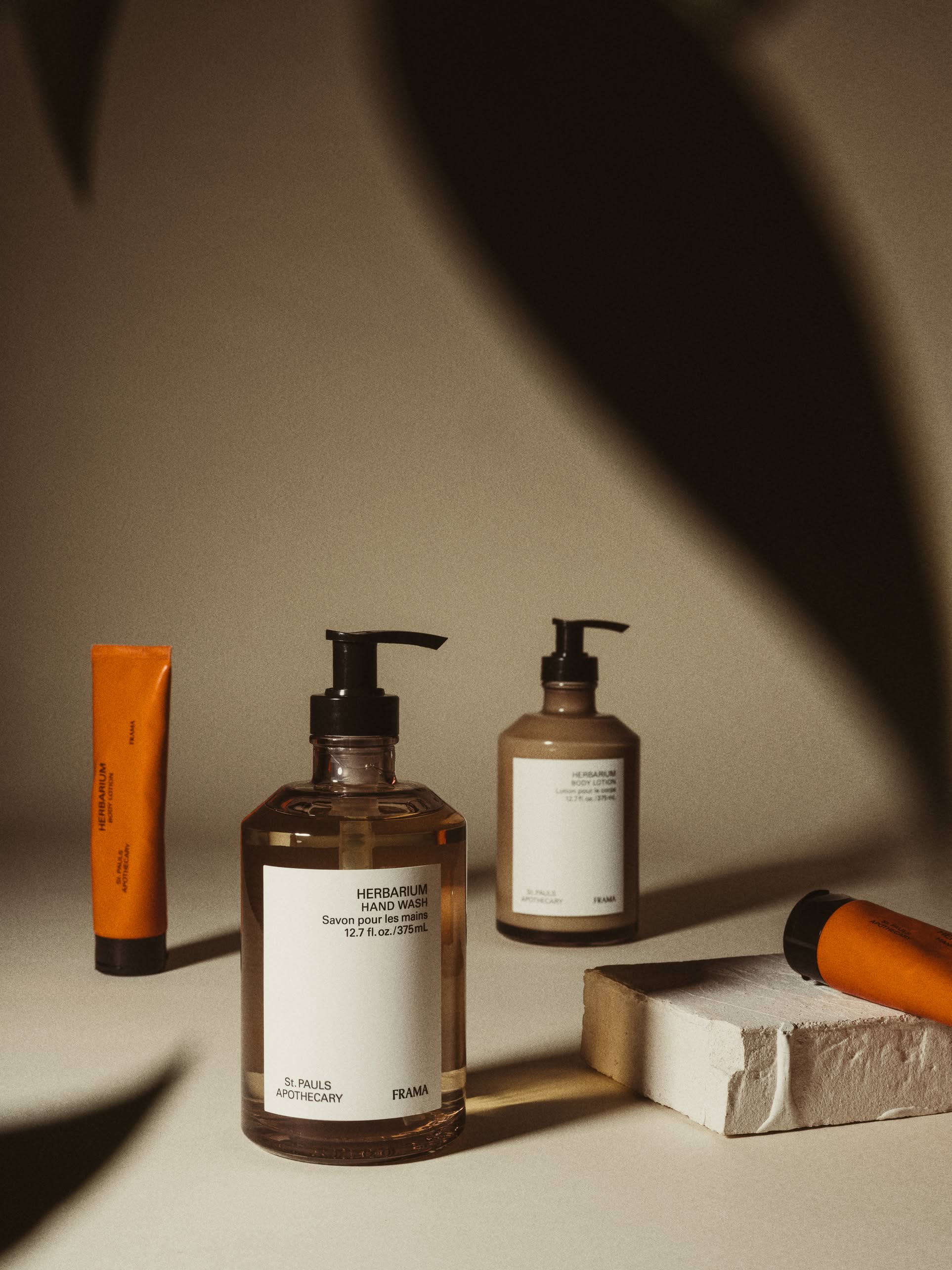 Minimalist photo of FRAMA Hand Wash, 375ml pump bottles and orange tubes, arranged on a beige surface with dramatic shadows and leafy silhouettes in the foreground.