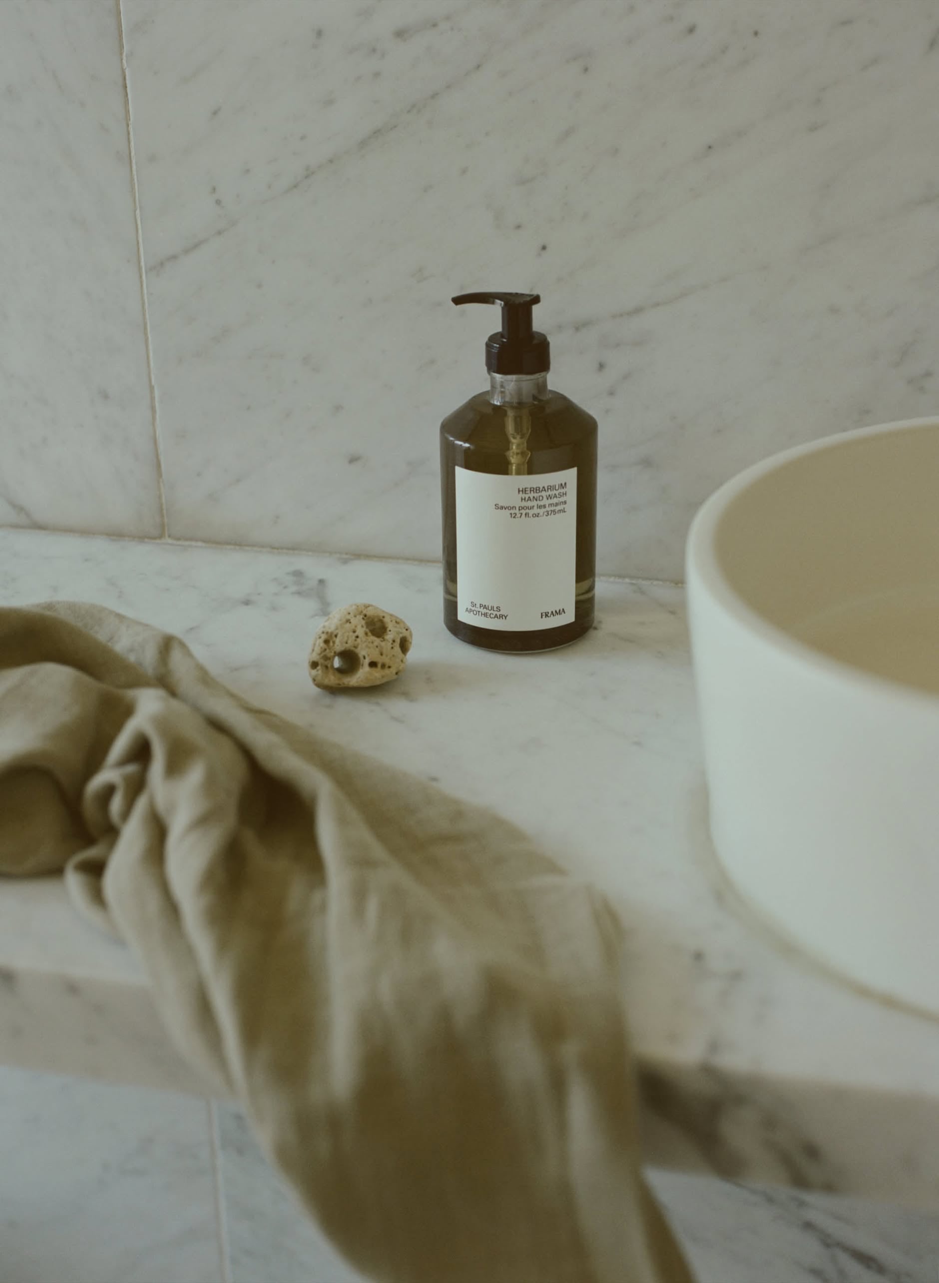 A 375ml brown pump bottle of FRAMA Hand Wash with organic ingredients sits on a marble bathroom countertop beside a light tan cloth, a small seashell, and a white round basin, against light gray marble tiles.