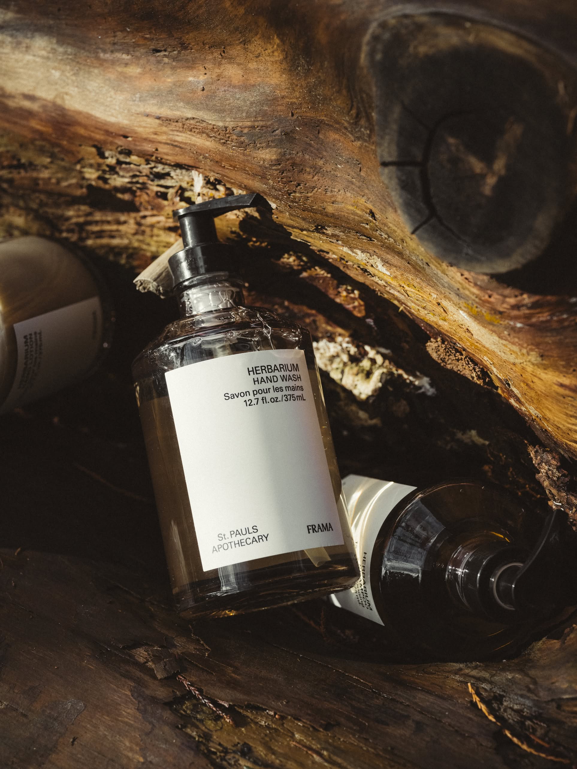 A bottle of FRAMA Hand Wash, 375ml, by FRAMA with a pump top sits inside a hollow tree trunk, with another bottle partially visible beside it in natural sunlight.
