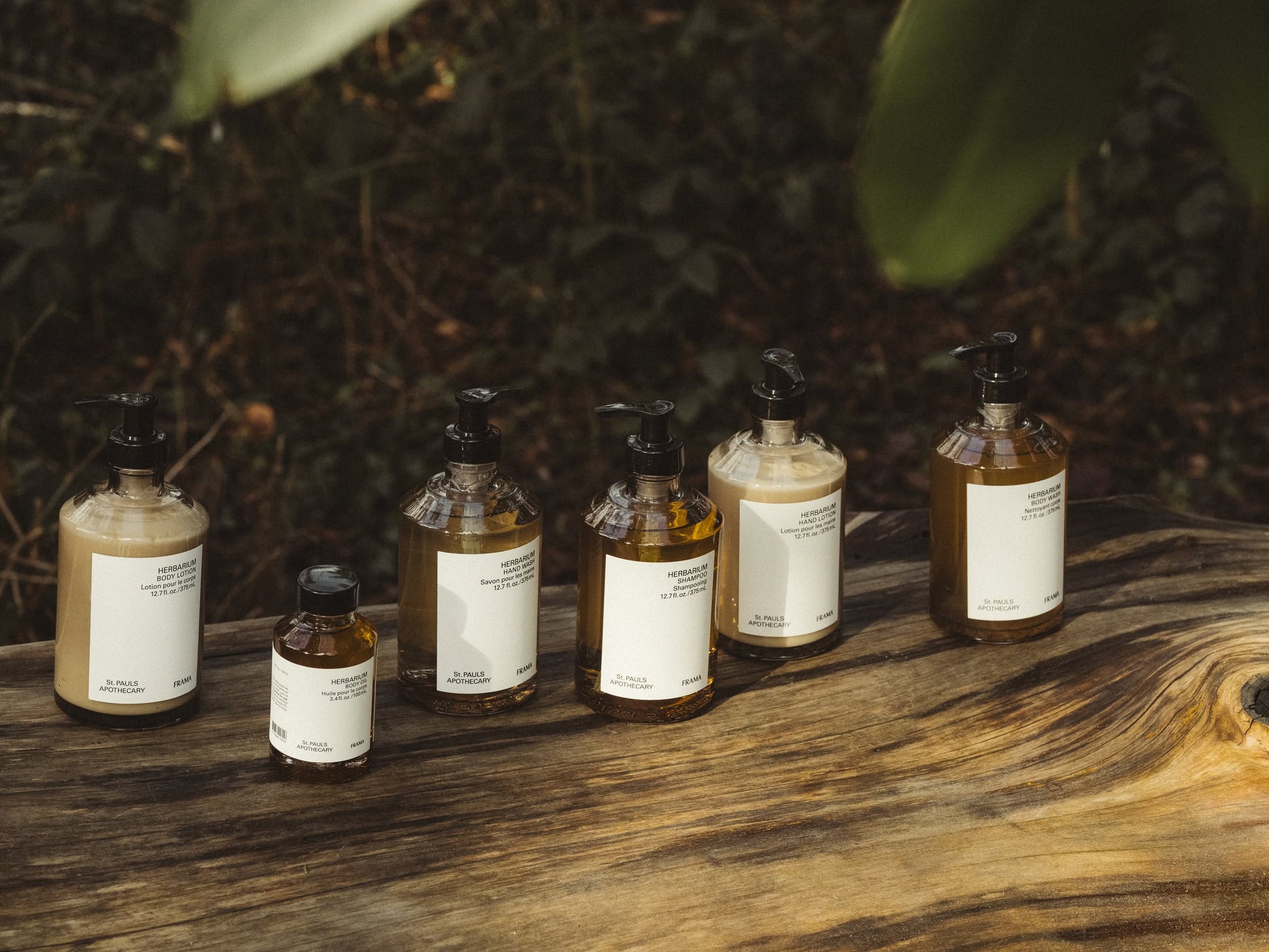 Six FRAMA Hand Wash 375ml bottles with white labels and black pump tops are arranged on a rustic outdoor wooden surface. The scene features organic ingredients, sandalwood scent, and softly blurred greenery in the background.