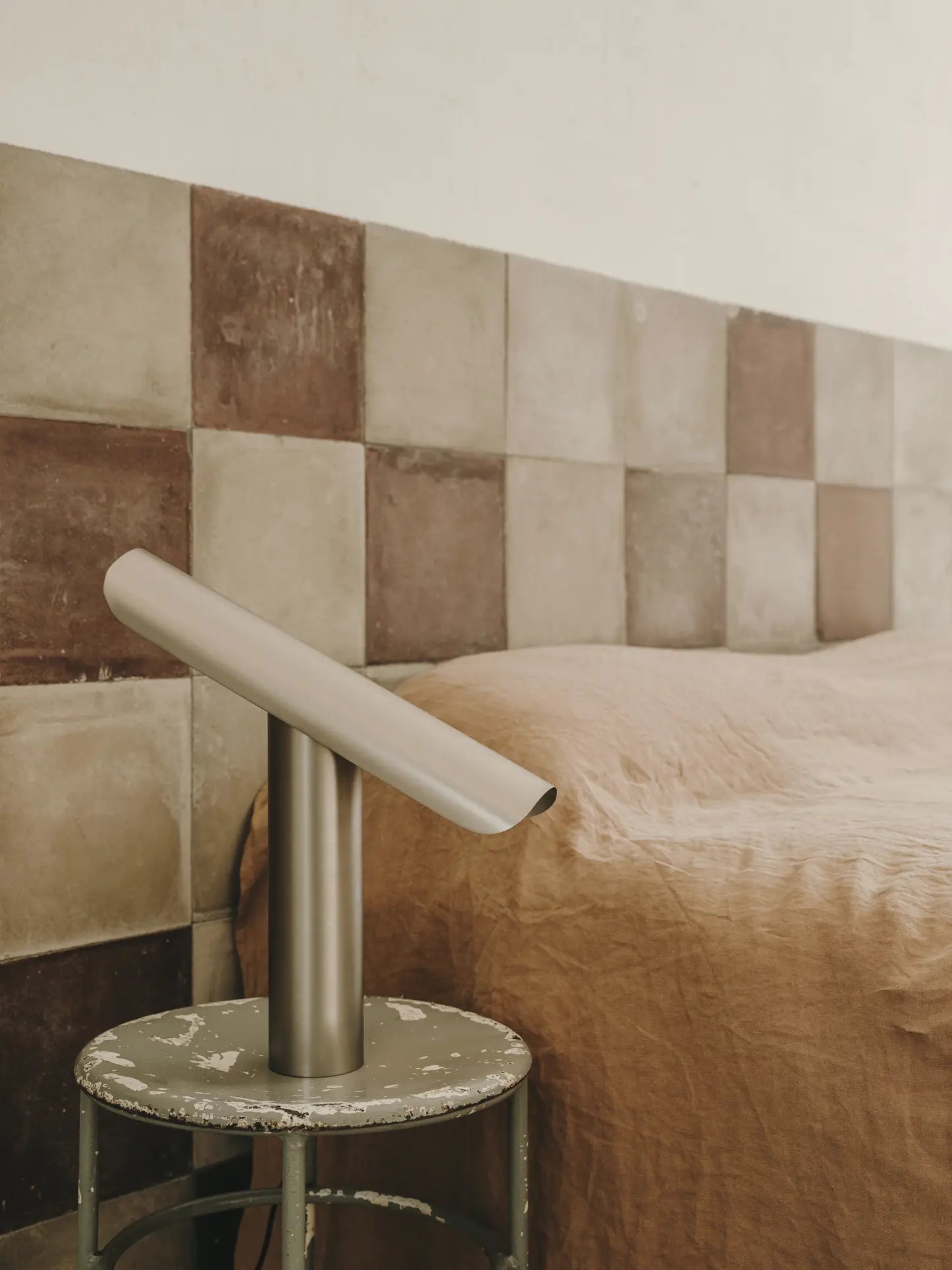 A minimalist bedroom features a beige and brown checkered tile headboard, a bed with a tan blanket, and the FRAMA T-Lamp Table Lamp by FRAMA on a round, weathered side table.