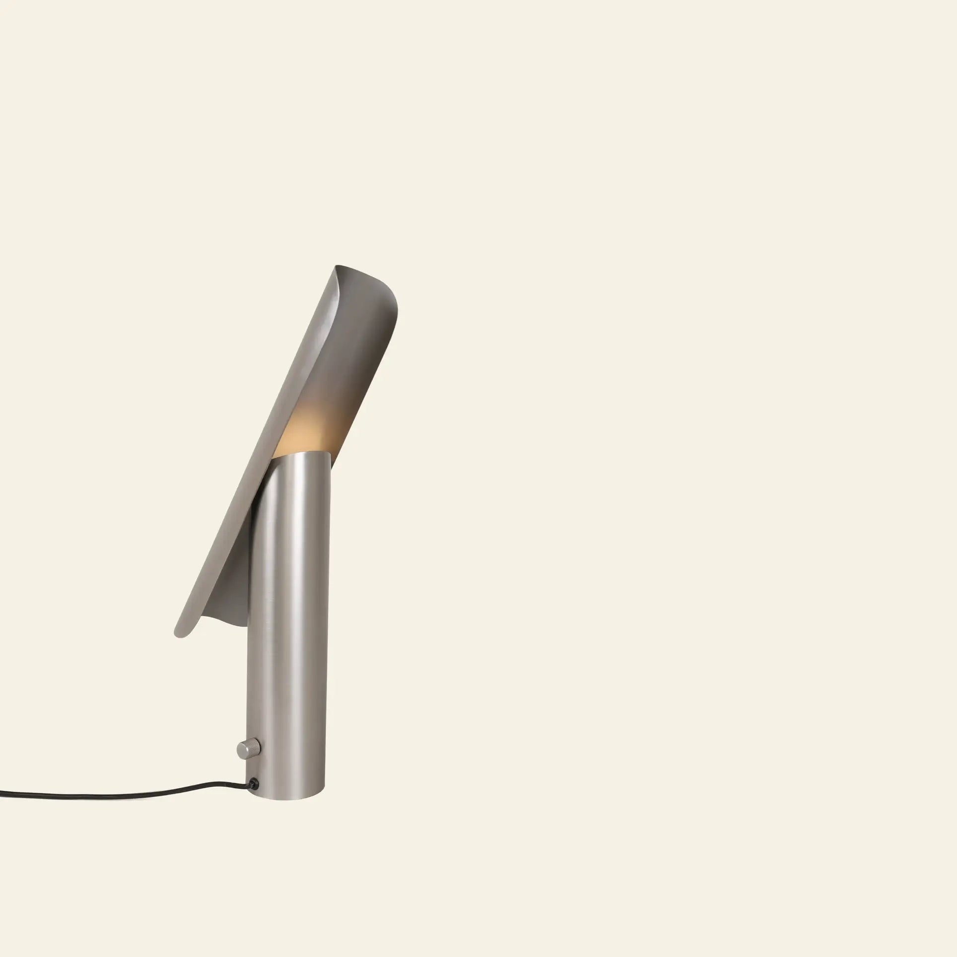 The FRAMA T-Lamp Table Lamp by FRAMA features a modern minimalist geometric design with a curved metallic shade and cylindrical base, emitting warm light. This contemporary desk lamp is plugged in and displayed against a light beige background.