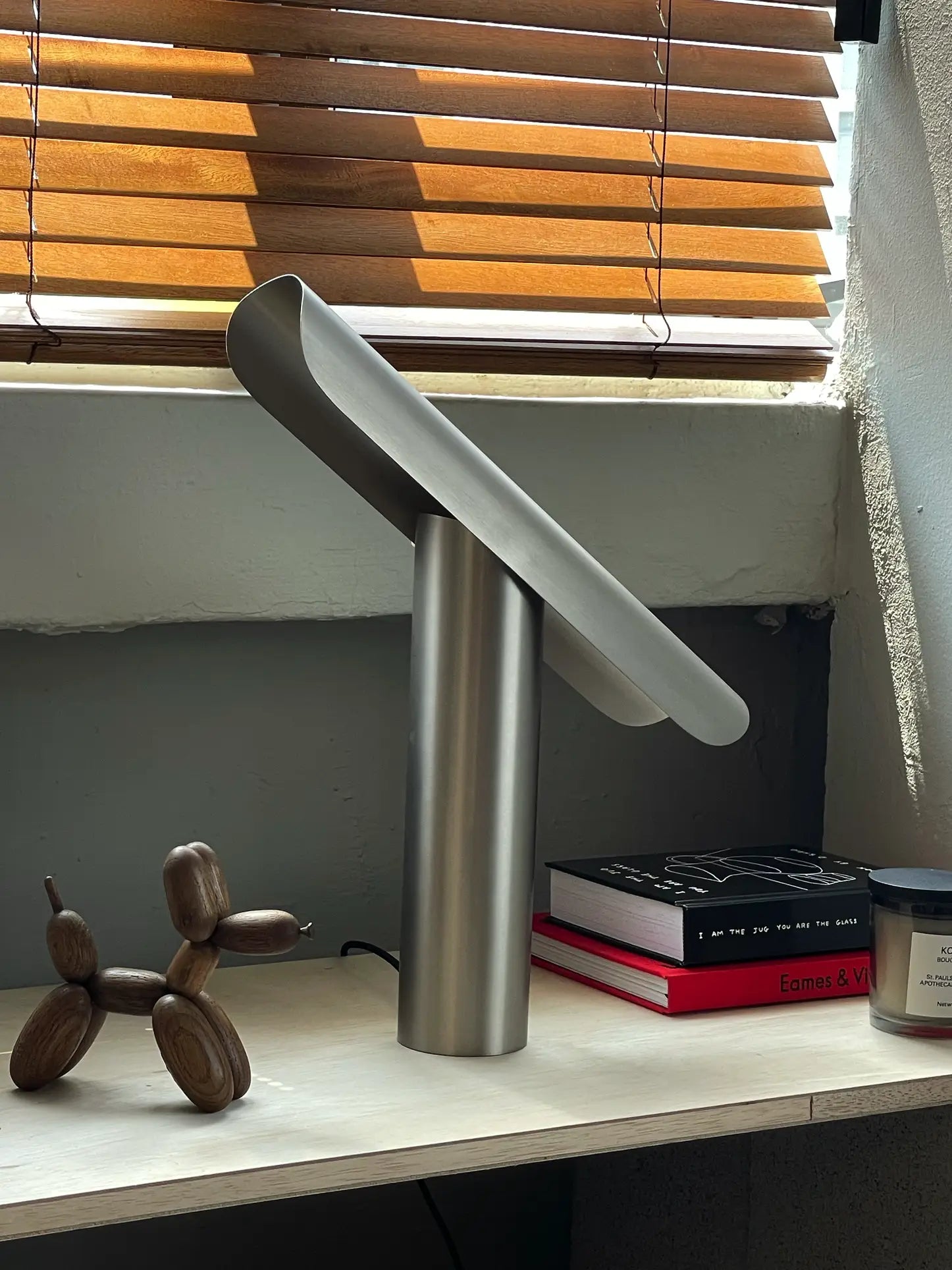 The FRAMA T-Lamp Table Lamp with an oval shade rests on a desk by wooden blinds, alongside a wooden balloon dog sculpture, two stacked books, and a red notebook as sunlight streams through the window.