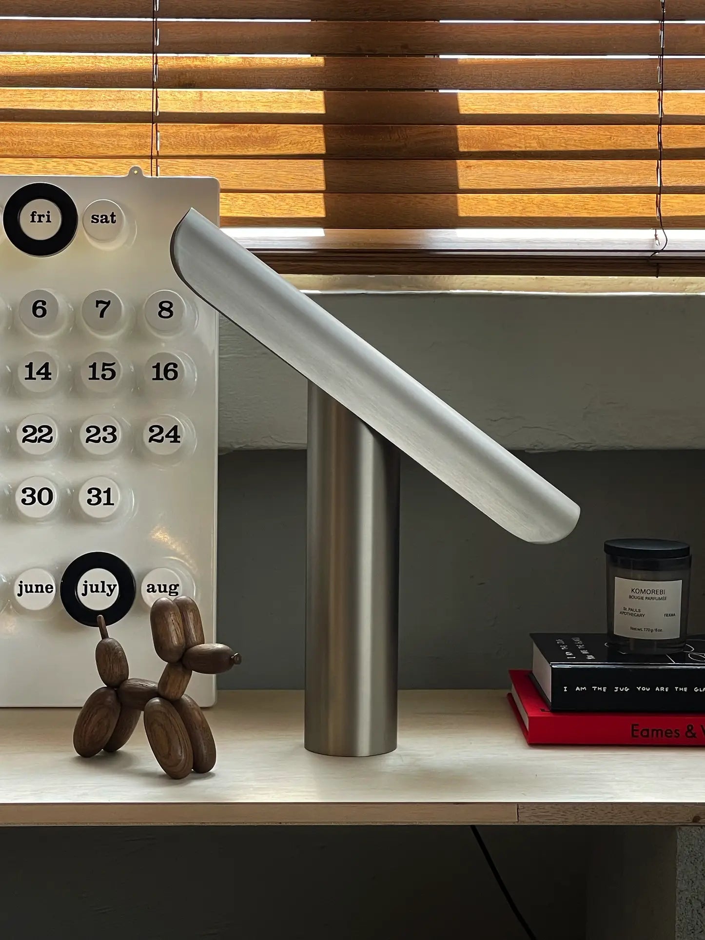 A minimalist desk features the FRAMA T-Lamp Table Lamp by FRAMA, a sculptural balloon dog figurine, a candle, books, and a bold black-and-white calendar beneath wooden blinds with sunlight streaming through.