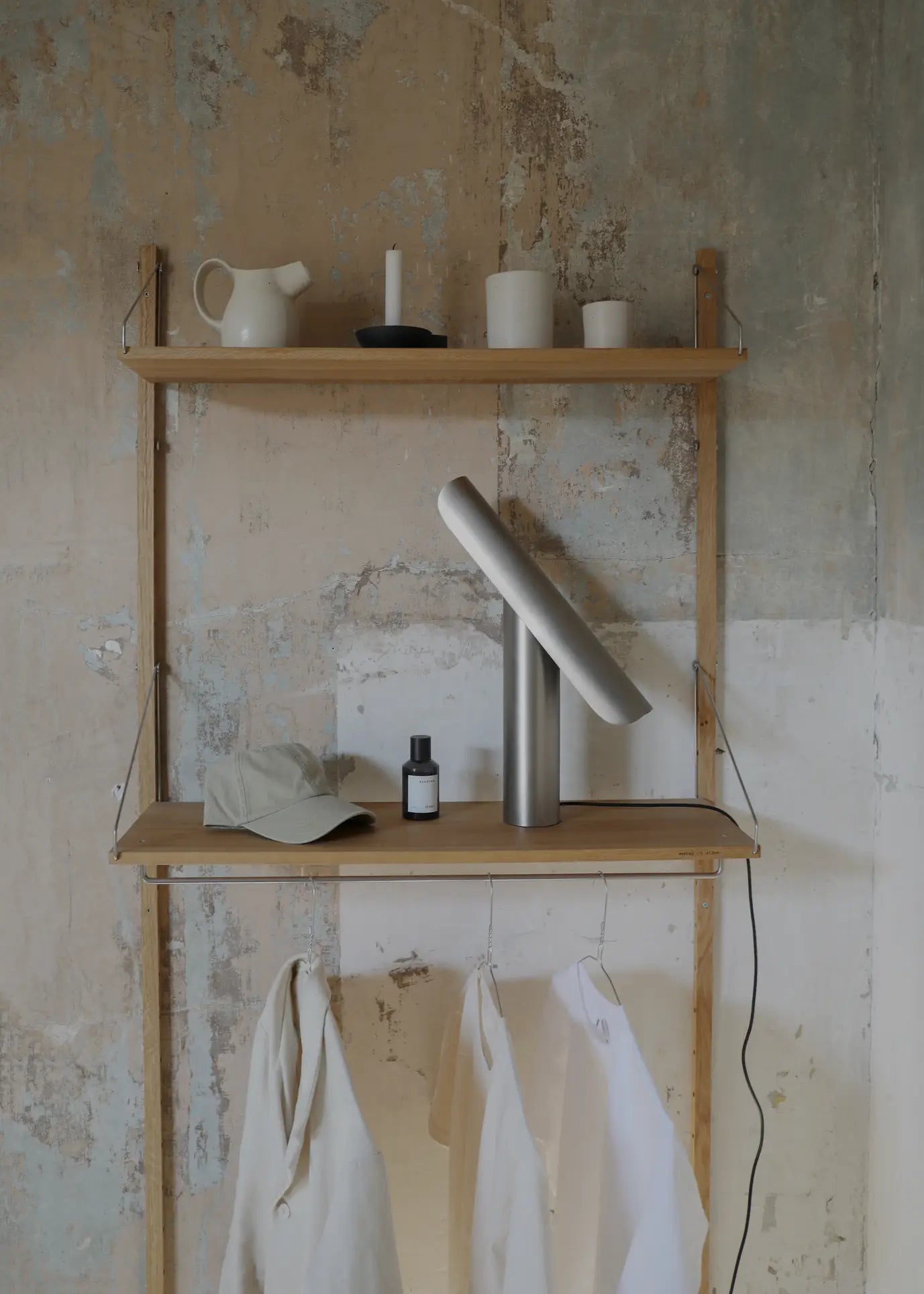A minimalist wooden shelf unit against a textured wall holds the FRAMA T-Lamp Table Lamp, a hat, a small bottle, and hanging white garments. The top shelf displays a pitcher, candle, and cups with the lamp’s power cord draped over the side.