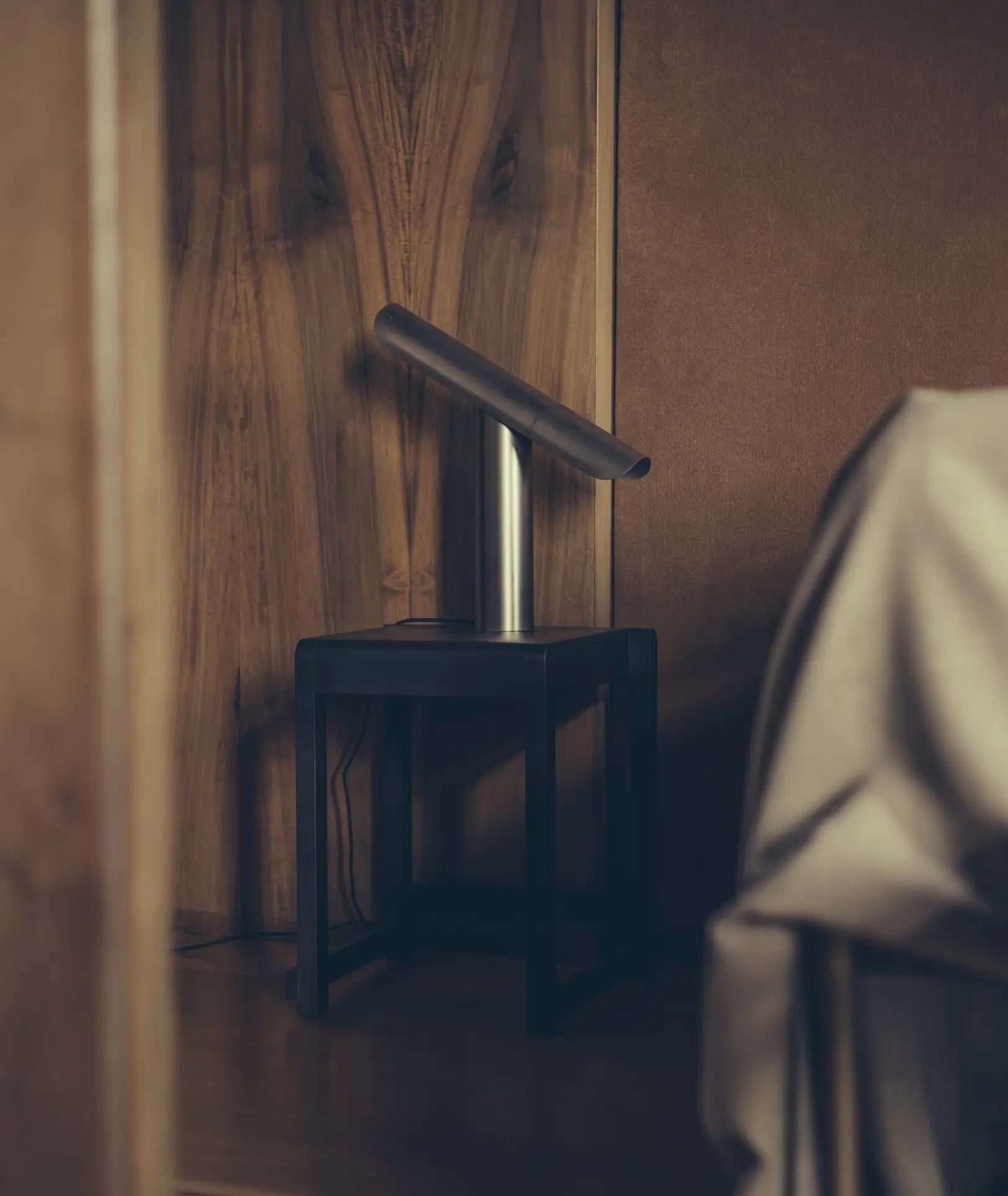 The FRAMA T-Lamp Table Lamp by FRAMA, featuring a minimalist geometric design and cylindrical shade, sits on a black side table in a cozy, dimly lit wood-paneled room with a beige cloth draped nearby.