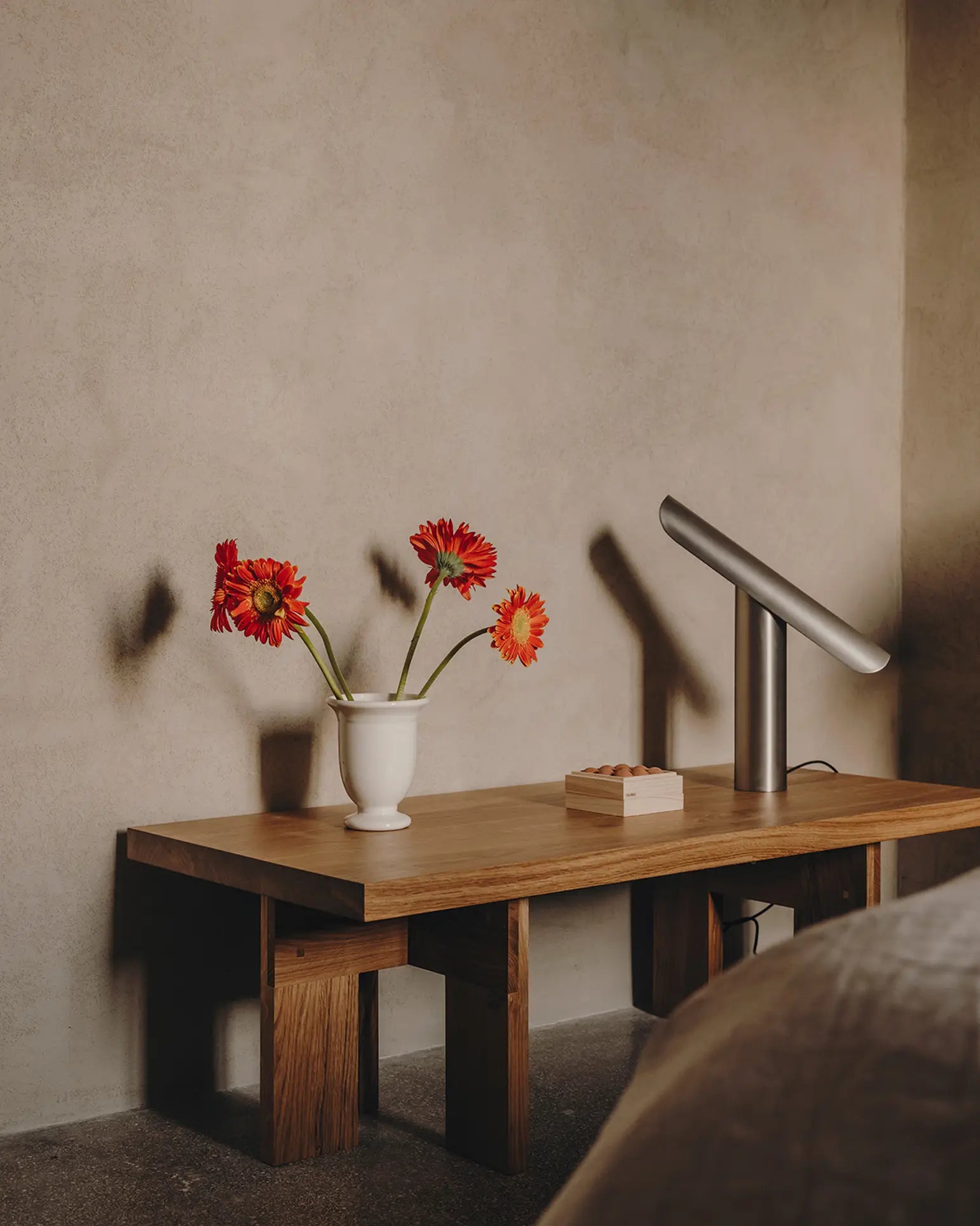 A wooden bedside table with a white vase of three red flowers, the FRAMA T-Lamp Table Lamp by FRAMA, and a closed book sits against a textured beige wall. Part of a bed is visible in the foreground.