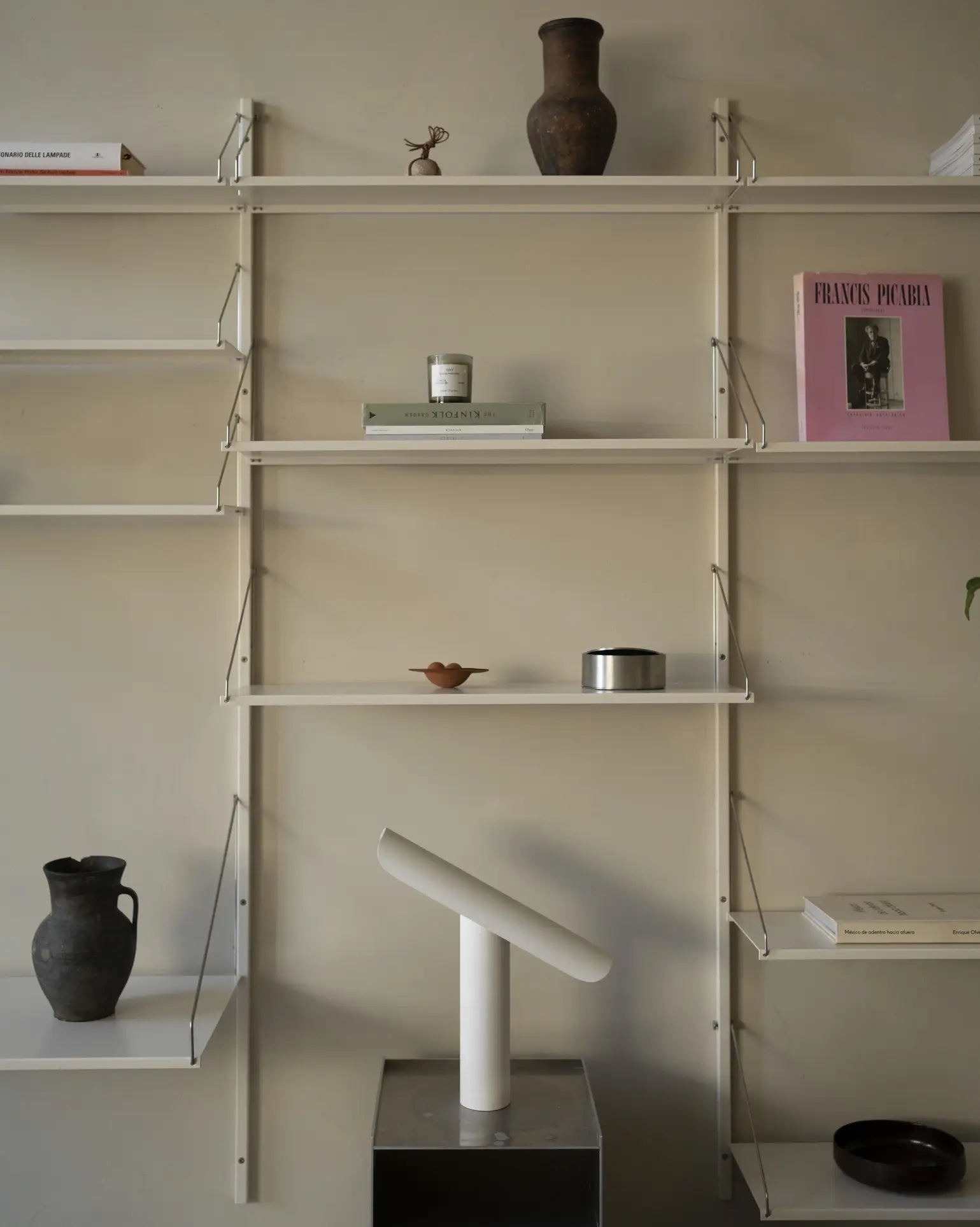 Minimalist wall-mounted shelves display books, a pink-framed photo, ceramic vases, and decor items. Below the shelves sits the FRAMA T-Lamp Table Lamp by FRAMA, featuring a contemporary geometric design on a gray pedestal.