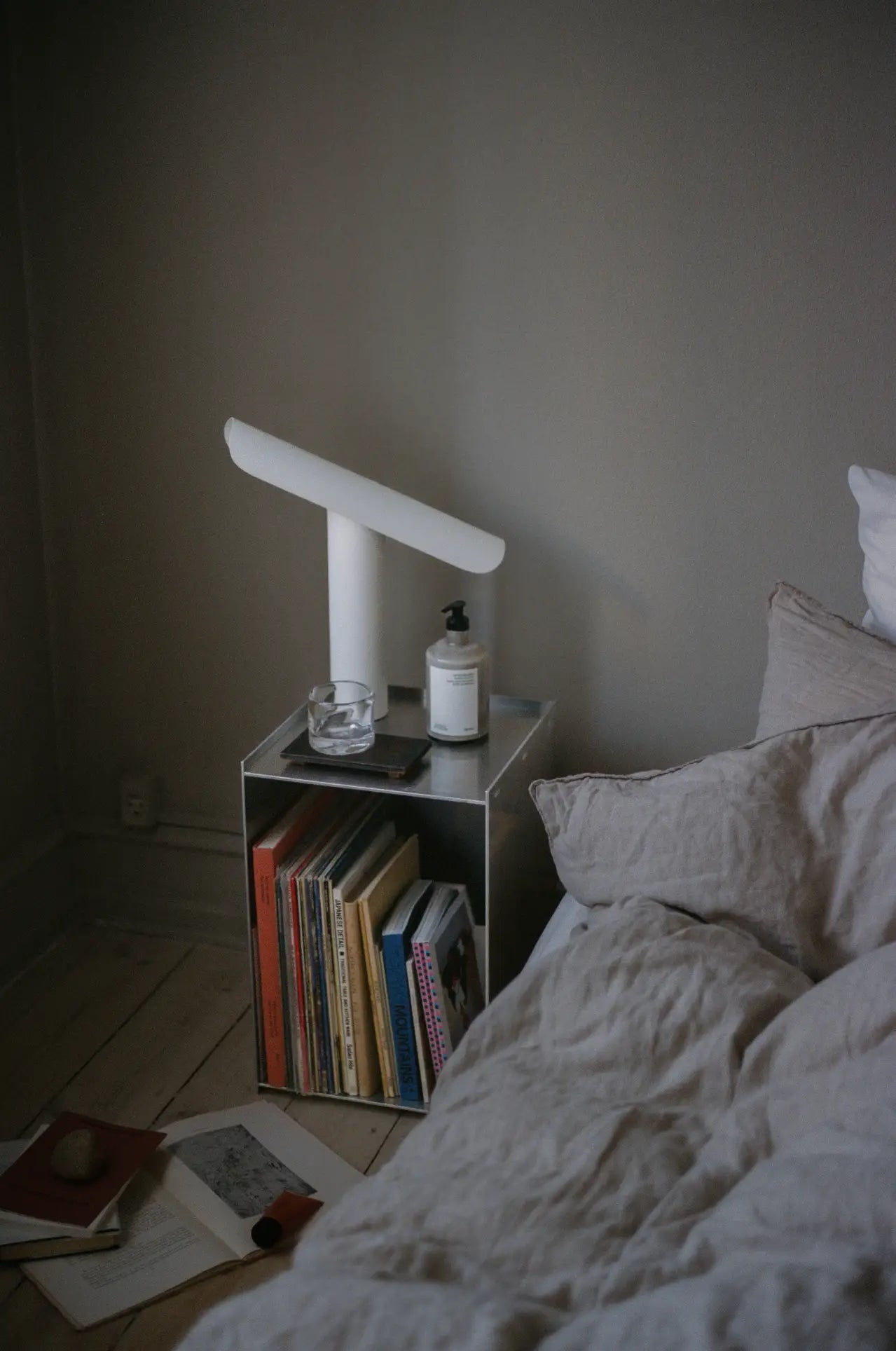 A cozy bedroom features an unmade bed, a small nightstand with the FRAMA T-Lamp Table Lamp by FRAMA, a candle, and a bottle. Several books and magazines are neatly stacked below, with an open notebook and pen on the floor.
