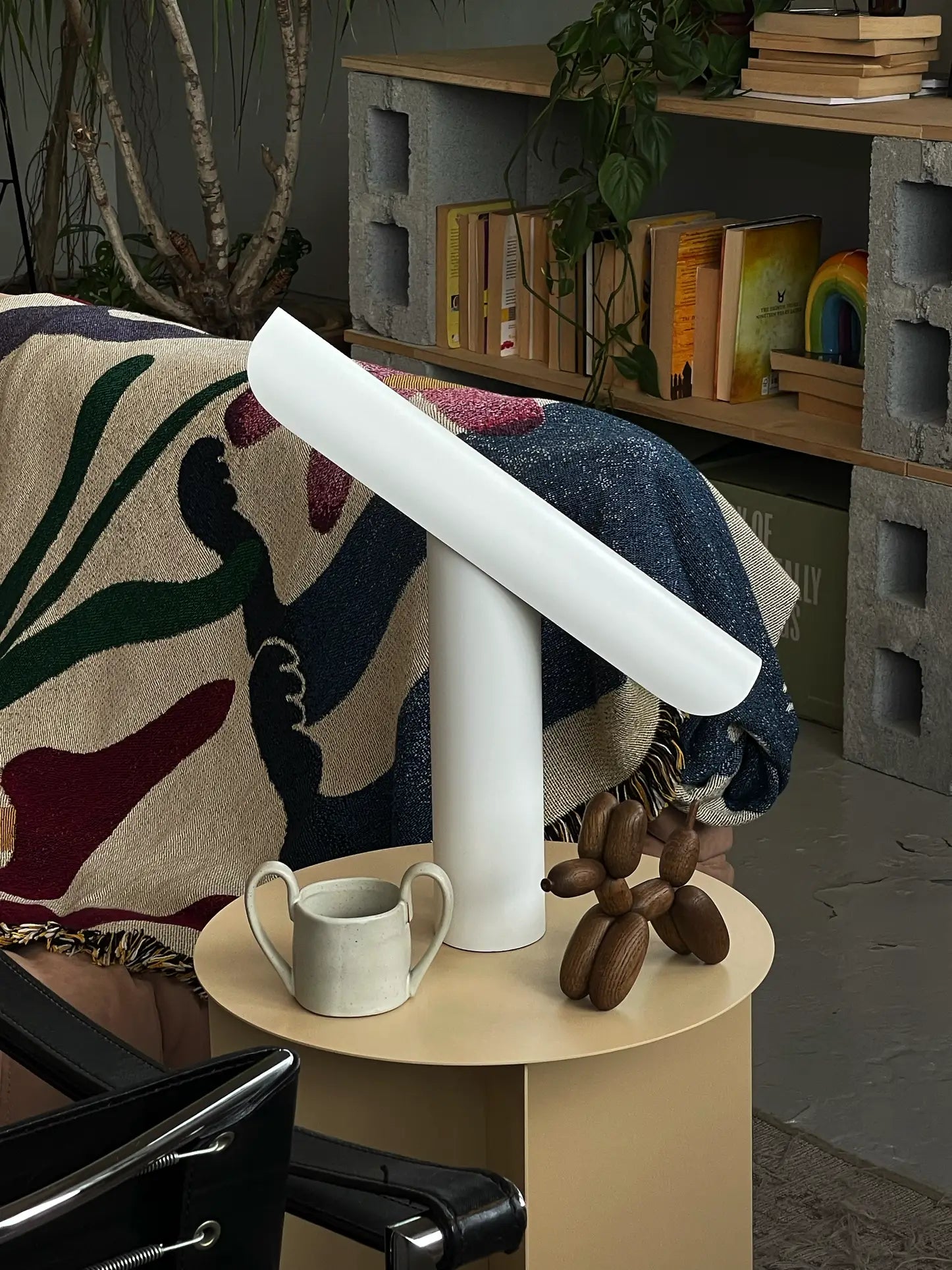 A modern living room features a beige side table with the FRAMA T-Lamp Table Lamp, a small cream teapot, and a wooden dog figurine. Behind is a sofa with an abstract blanket, while books and plants sit on the shelf.