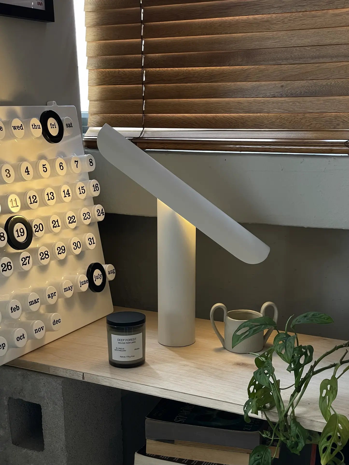 A FRAMA T-Lamp Table Lamp, a calendar, a potted plant, a candle, and a mug rest on a wooden shelf by closed blinds, creating an organized and cozy workspace.