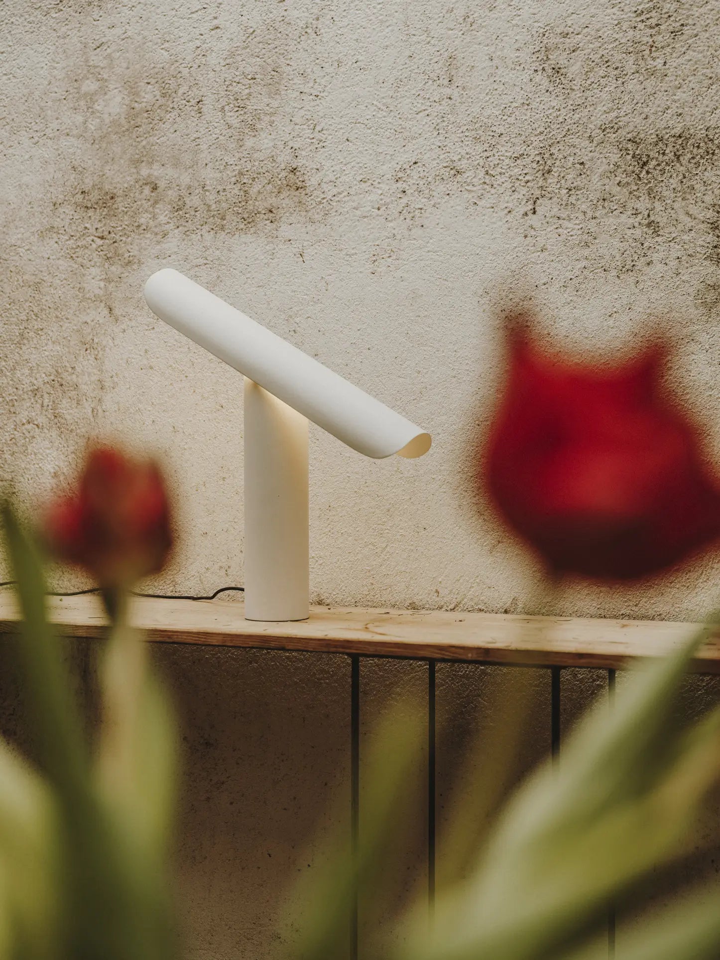 The FRAMA T-Lamp Table Lamp by FRAMA, featuring a minimalist geometric design and angled cylindrical shade, sits on a wooden surface against a textured wall with blurred red flowers in the foreground.