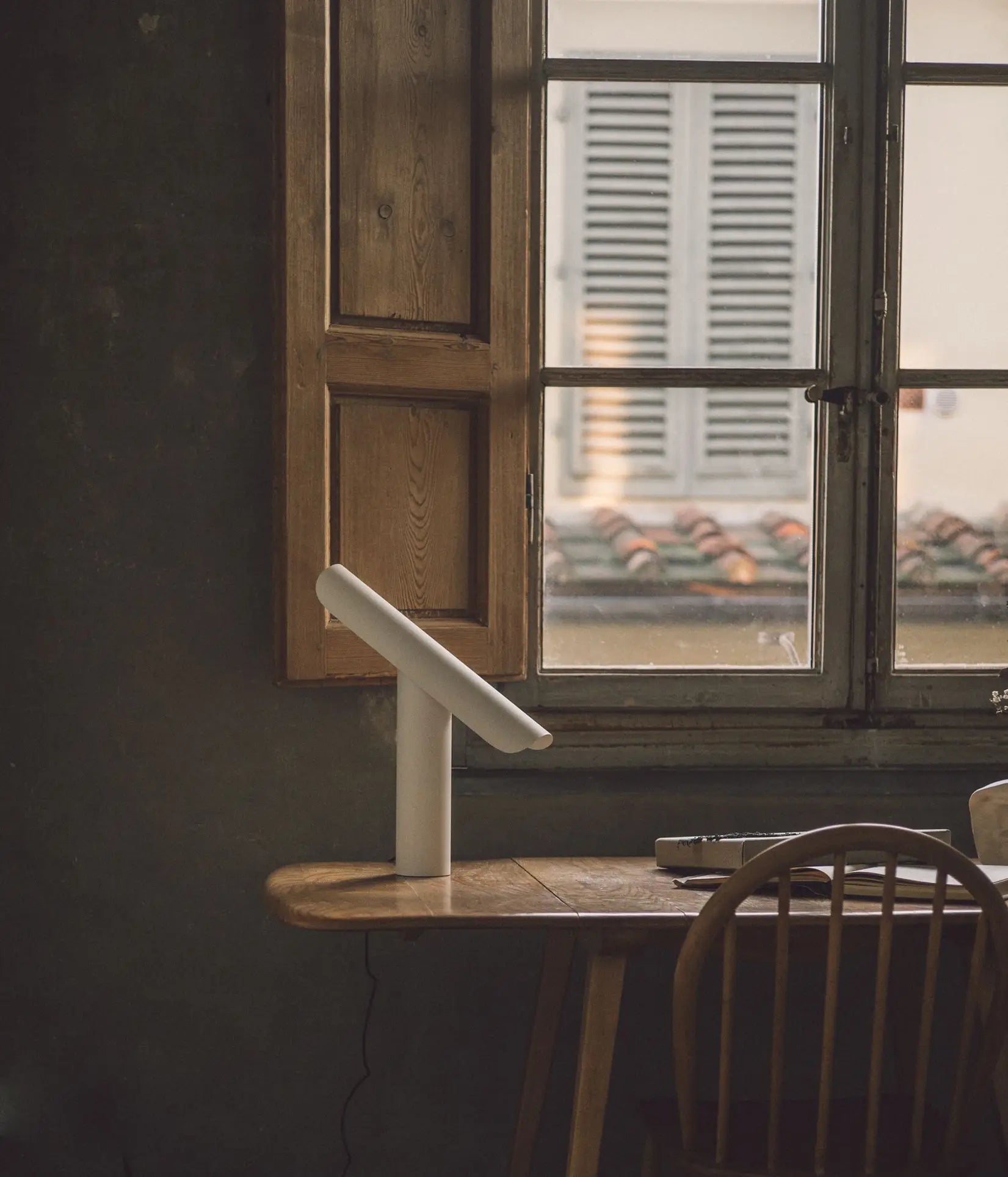 A wooden desk with a FRAMA T-Lamp Table Lamp by FRAMA sits by a window with wooden shutters, overlooking a building with tiled roofing and closed window shutters outside. A wooden chair is tucked beneath the desk.