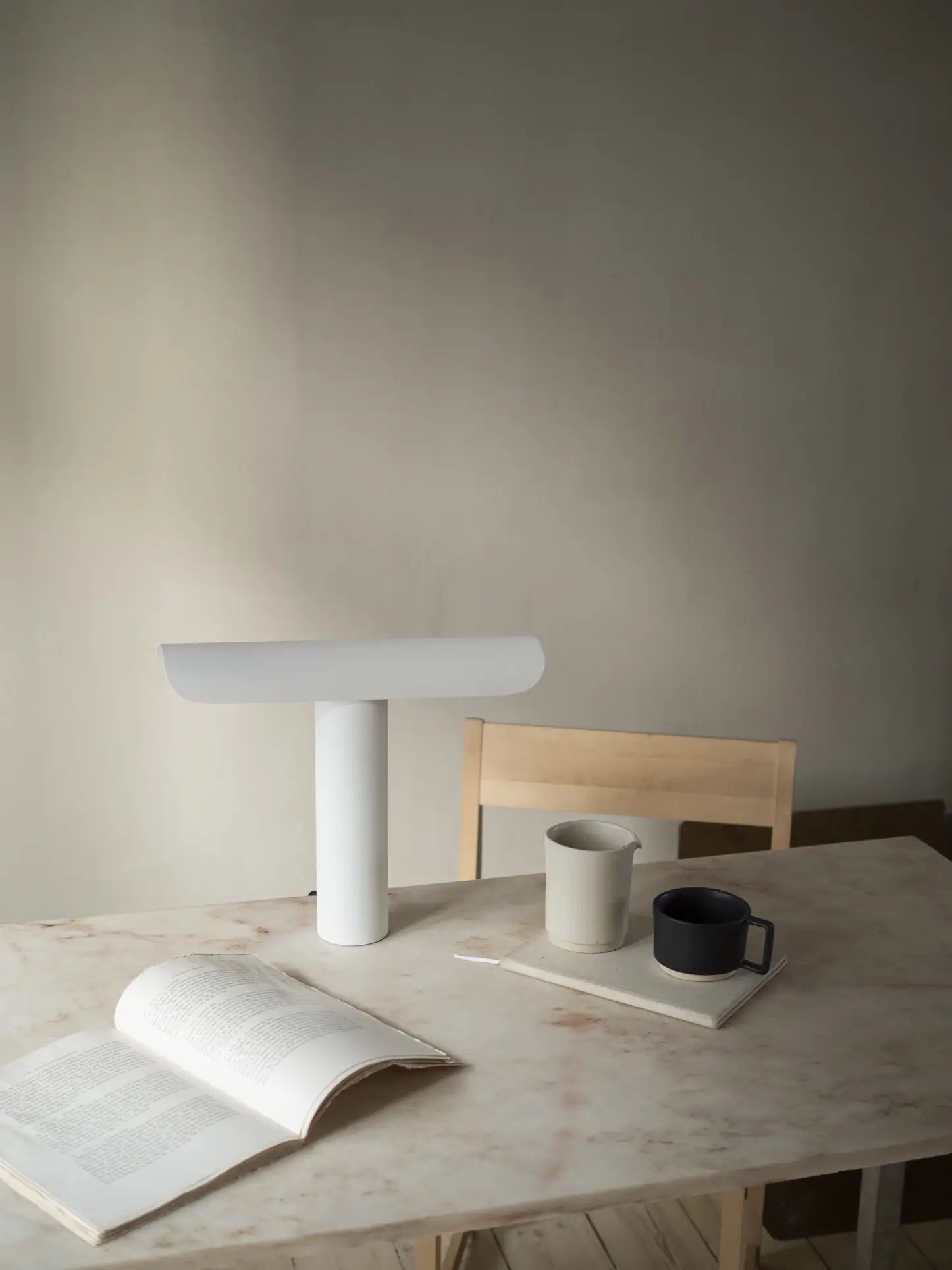 A minimalistic table with an open book, the FRAMA T-Lamp Table Lamp by FRAMA featuring a tilting mechanism, two mugs—one beige, one black—on a tray, and a light wooden chair, all set against a plain beige wall.