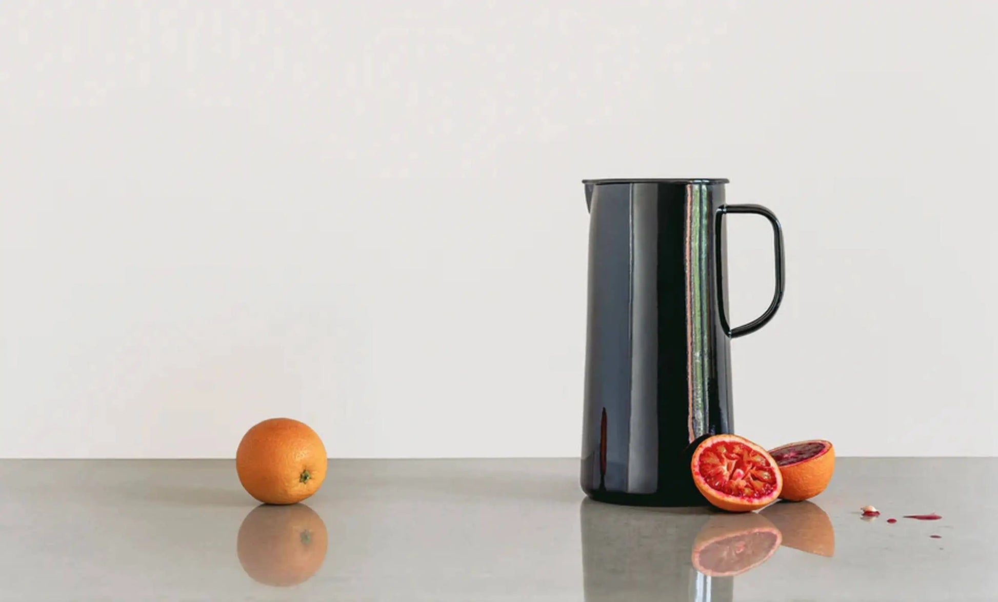 A whole orange, a halved blood orange, and a tall black Falcon Enamelware 3 Pint Jug by Falcon Enamelware sit on a reflective gray surface against a plain white background.