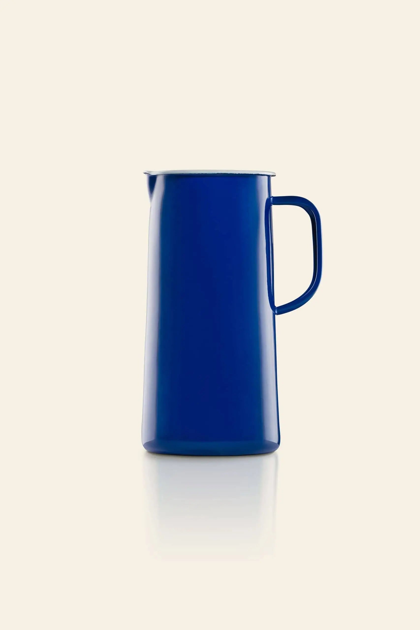 A tall, sleek Falcon Enamelware 3 Pint Jug in blue enamel with a handle stands centered on a plain light beige background. Its minimalist retro design features a slightly flared spout and its reflection appears below.