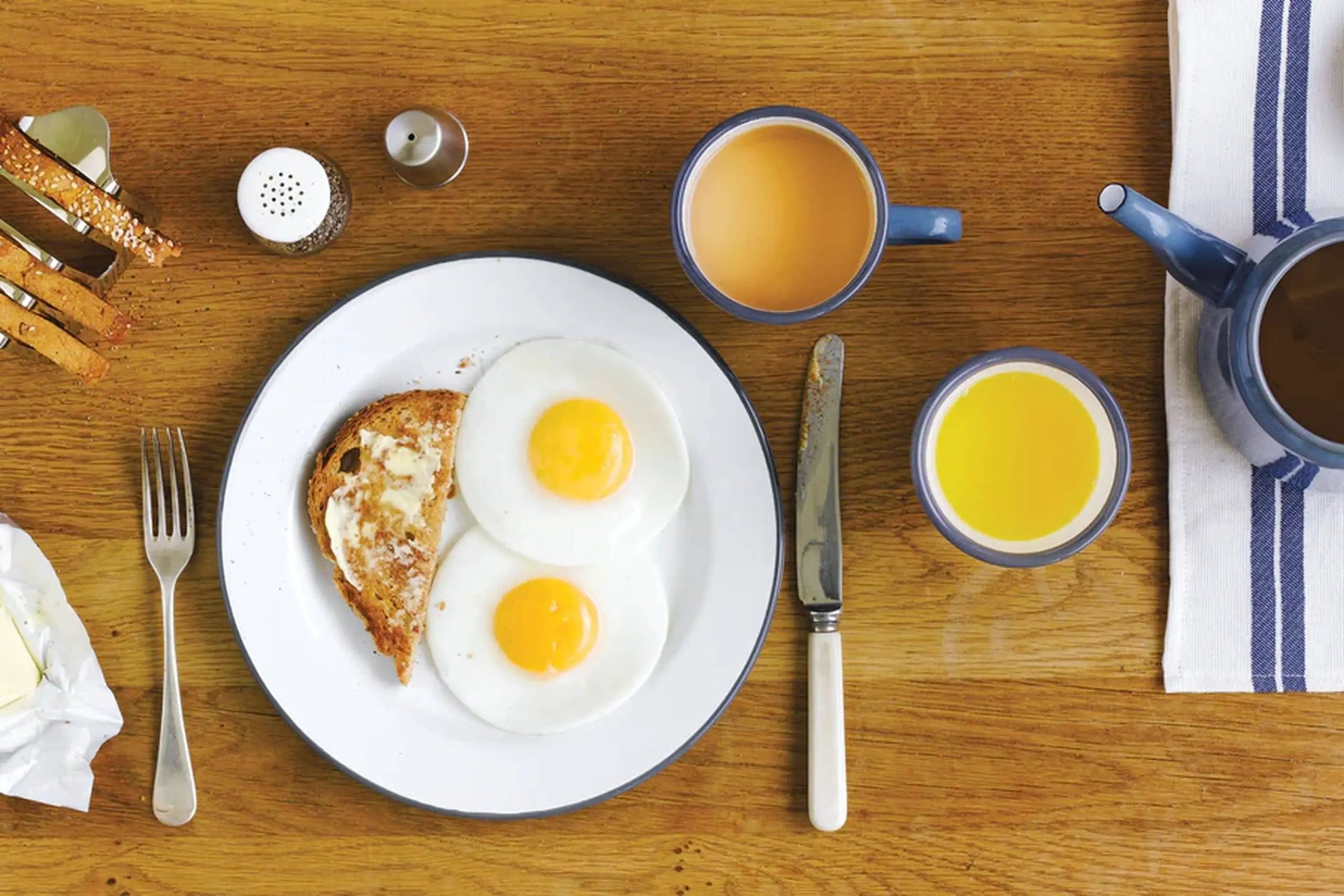 A breakfast plate with two sunny-side-up eggs and buttered toast sits beside a Falcon Enamelware Mug by Falcon Enamelware filled with coffee, plus orange juice, cutlery, and salt and pepper shakers on a wooden table for classic vintage style.