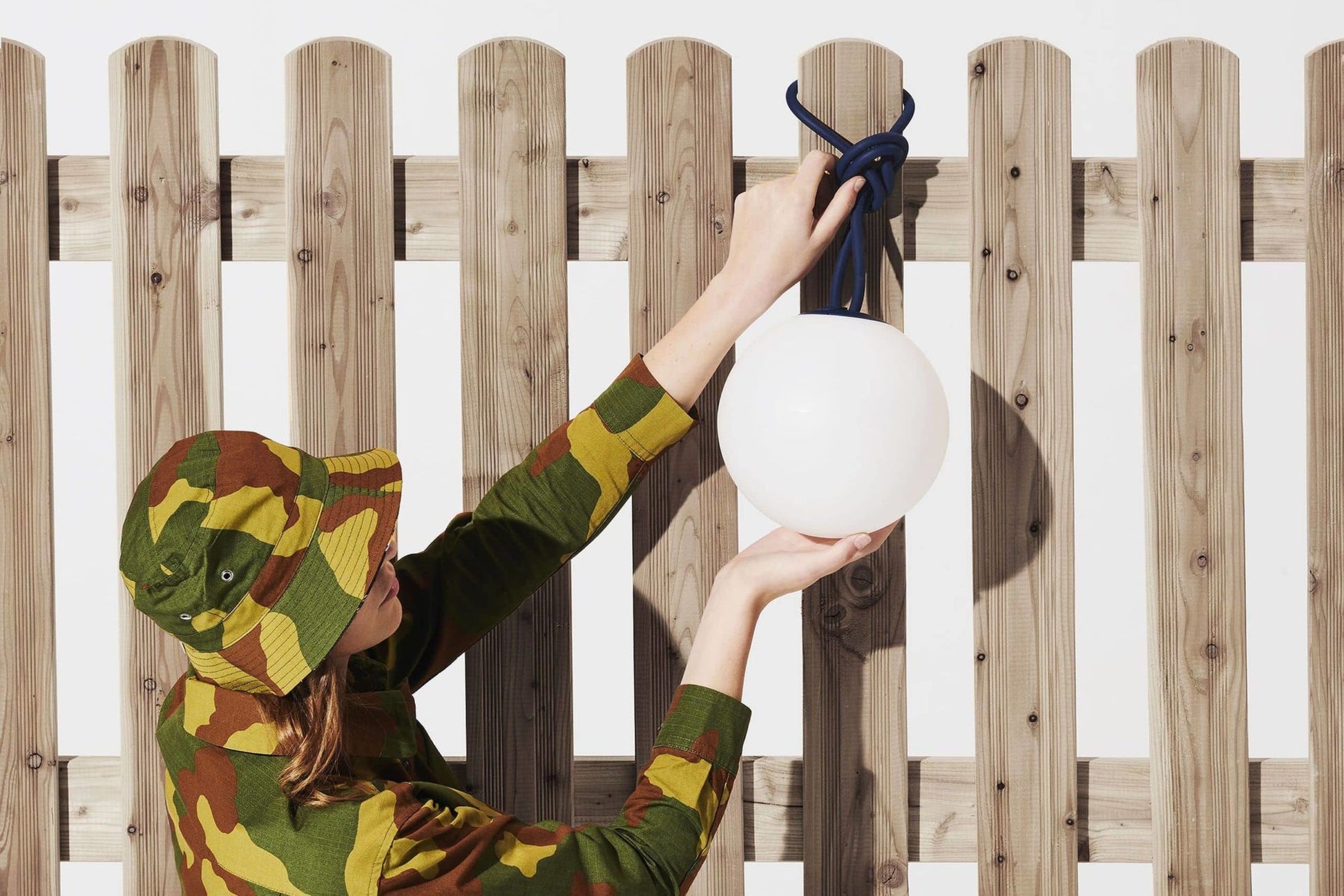A person in camouflage attire hangs the Fatboy Bolleke, a round white light by Fatboy, with a blue cord on a wooden fence, as sunlight creates modern shadows.