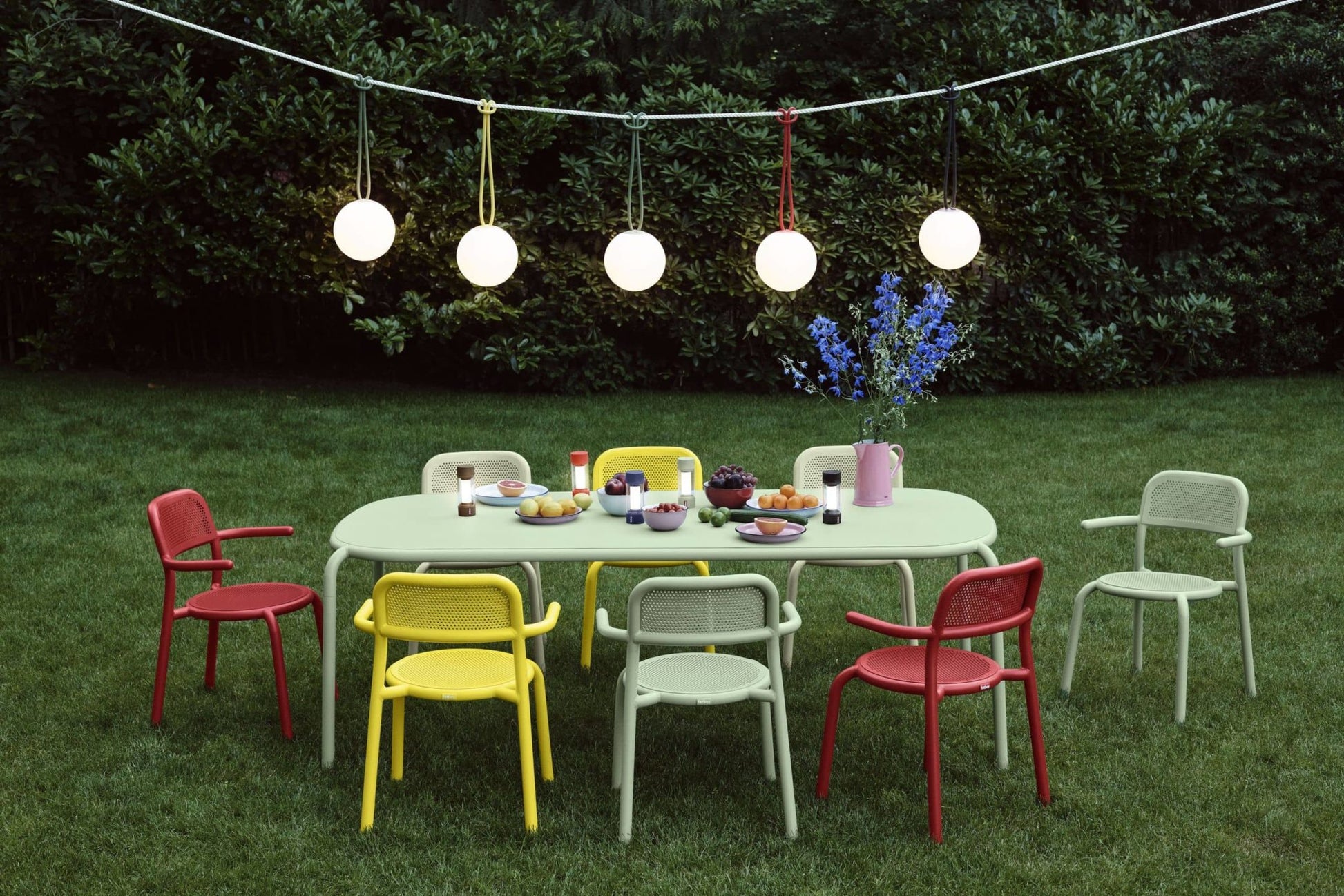 A modern outdoor dining set with colorful chairs, fruit, drinks, and flowers sits on grass beneath glowing Fatboy Bolleke lights, with lush green bushes in the background.