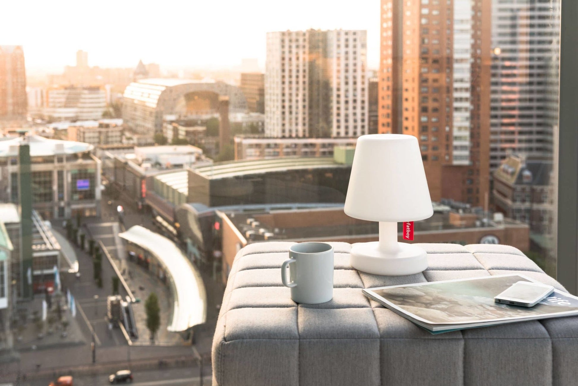 A Fatboy Edison the Petit lamp, a gray mug, a magazine, and a smartphone sit on an ottoman by a large window with sunset views of tall city buildings.