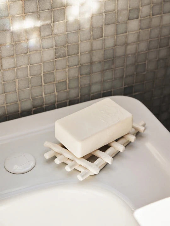 A Ferm Living Ceramic Soap Tray in off-white holds a white bar of soap atop a bathroom sink, with tiled wall tiles visible in the background.