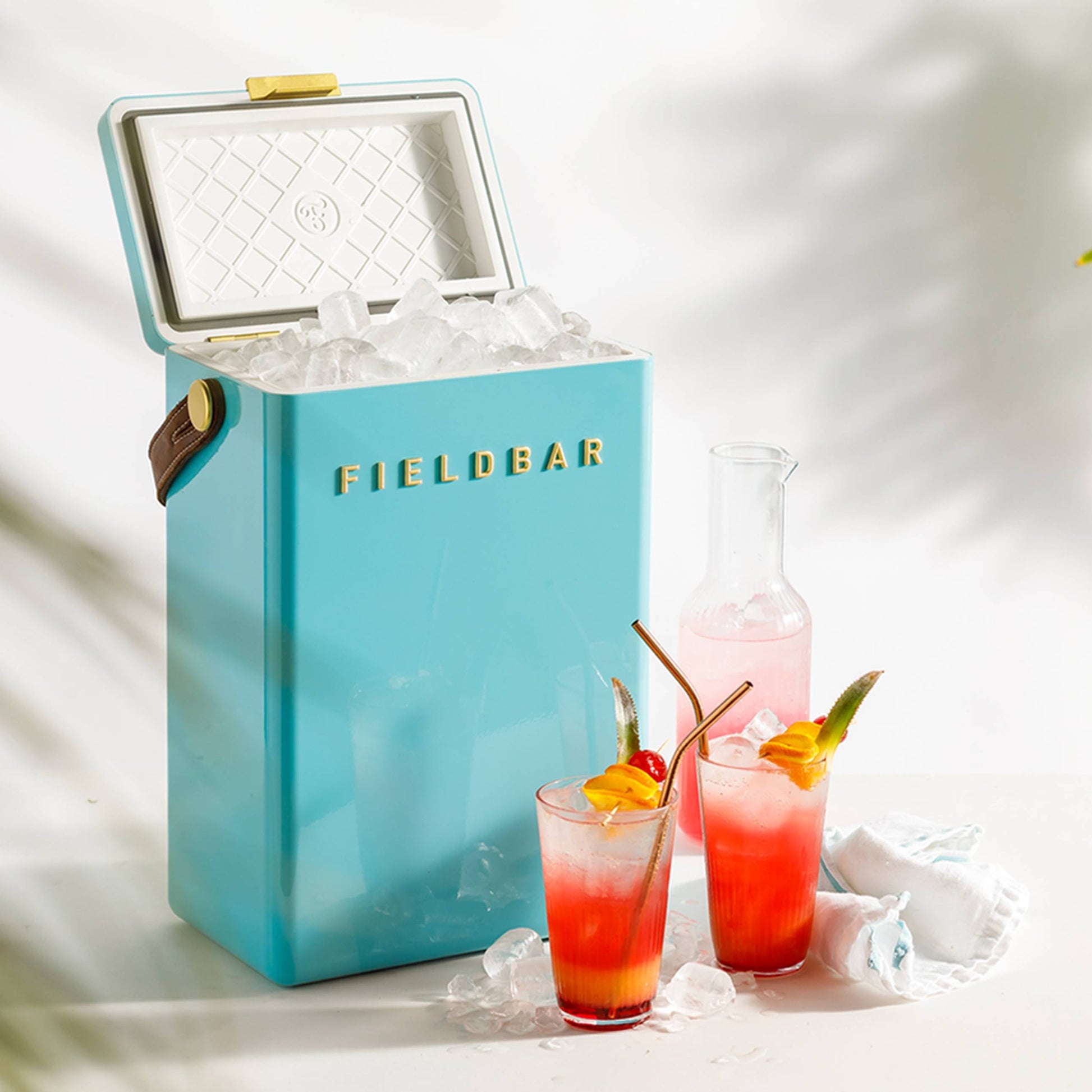 A blue Fieldbar Drinks Box by Fieldbar, filled with ice, sits beside two fruit-garnished tropical cocktails and a glass carafe on a white surface with scattered ice and a folded cloth—highlighting its top thermal performance.