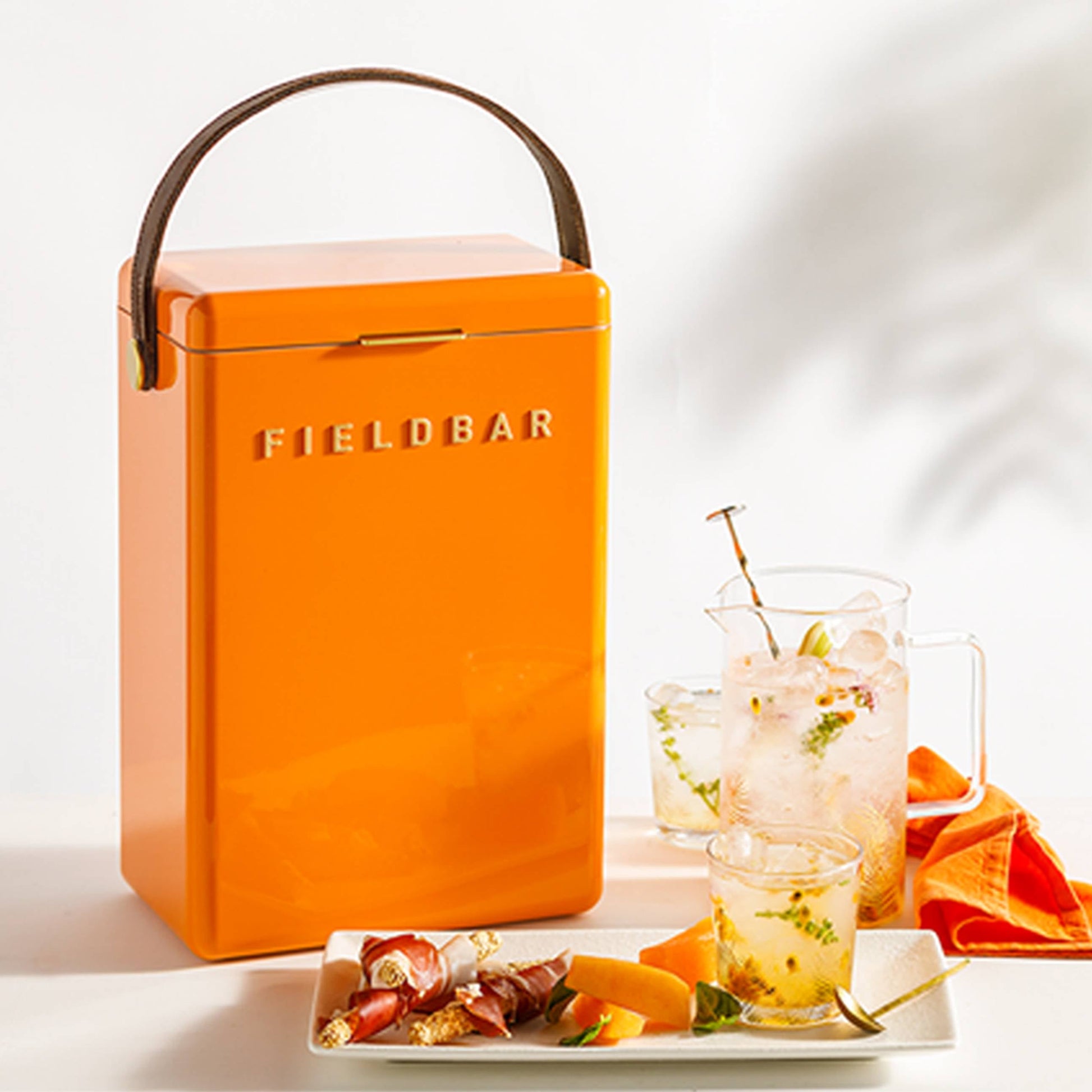 A bright orange, rectangular Fieldbar Drinks Box by Fieldbar sits beside appetizers, a glass of ice-filled herbal drink, a matching glass pitcher, and an orange napkin on a light surface.
