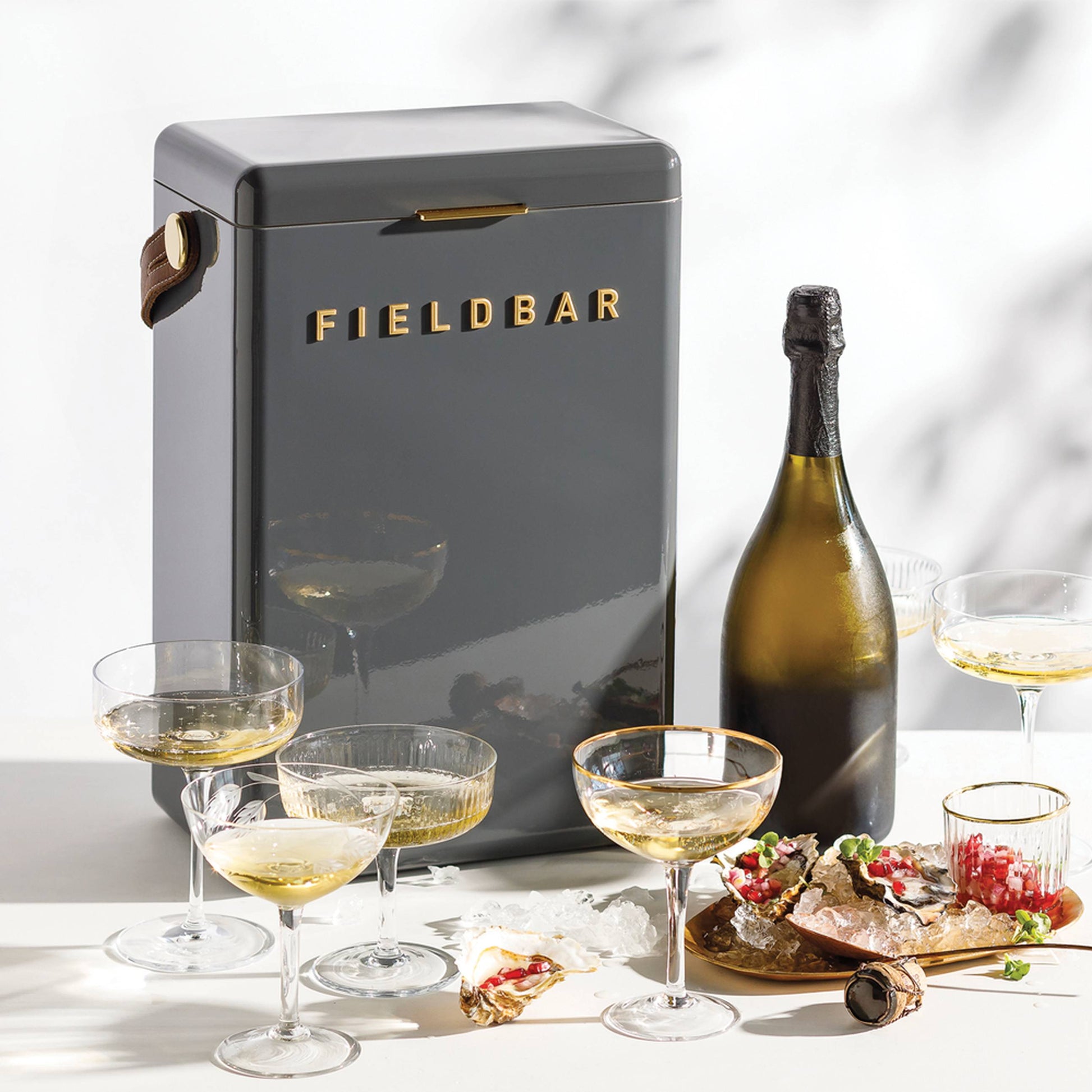 A sleek, dark gray Fieldbar Drinks Box by Fieldbar rests on a white surface next to sparkling wine, filled champagne glasses, ice cubes, and a plate of appetizers. Sunlight casts soft shadows in the background.
