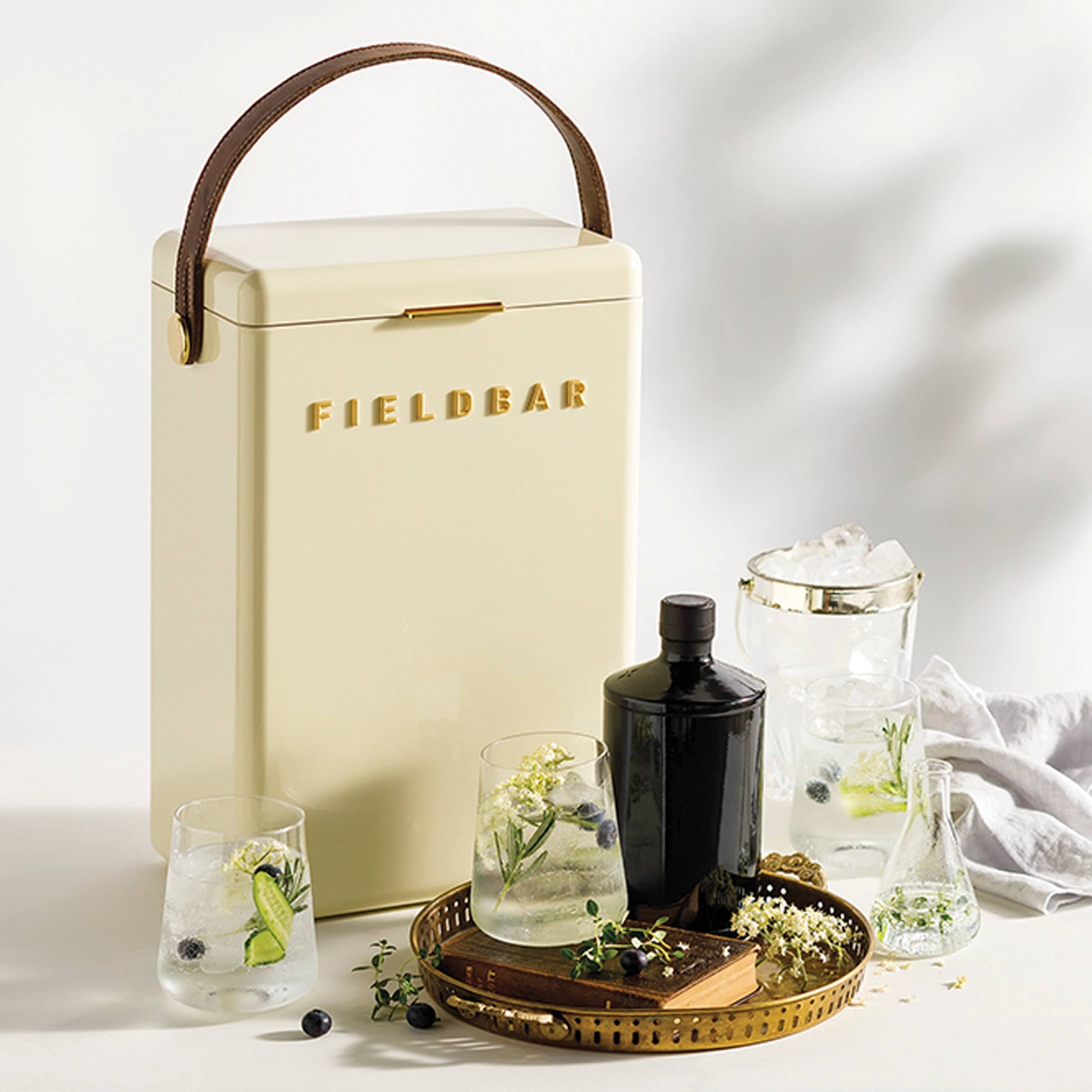 A cream-colored Fieldbar Drinks Box by Fieldbar with a brown handle sits beside glasses of iced drinks, a black bottle, and a gold tray on a white surface.