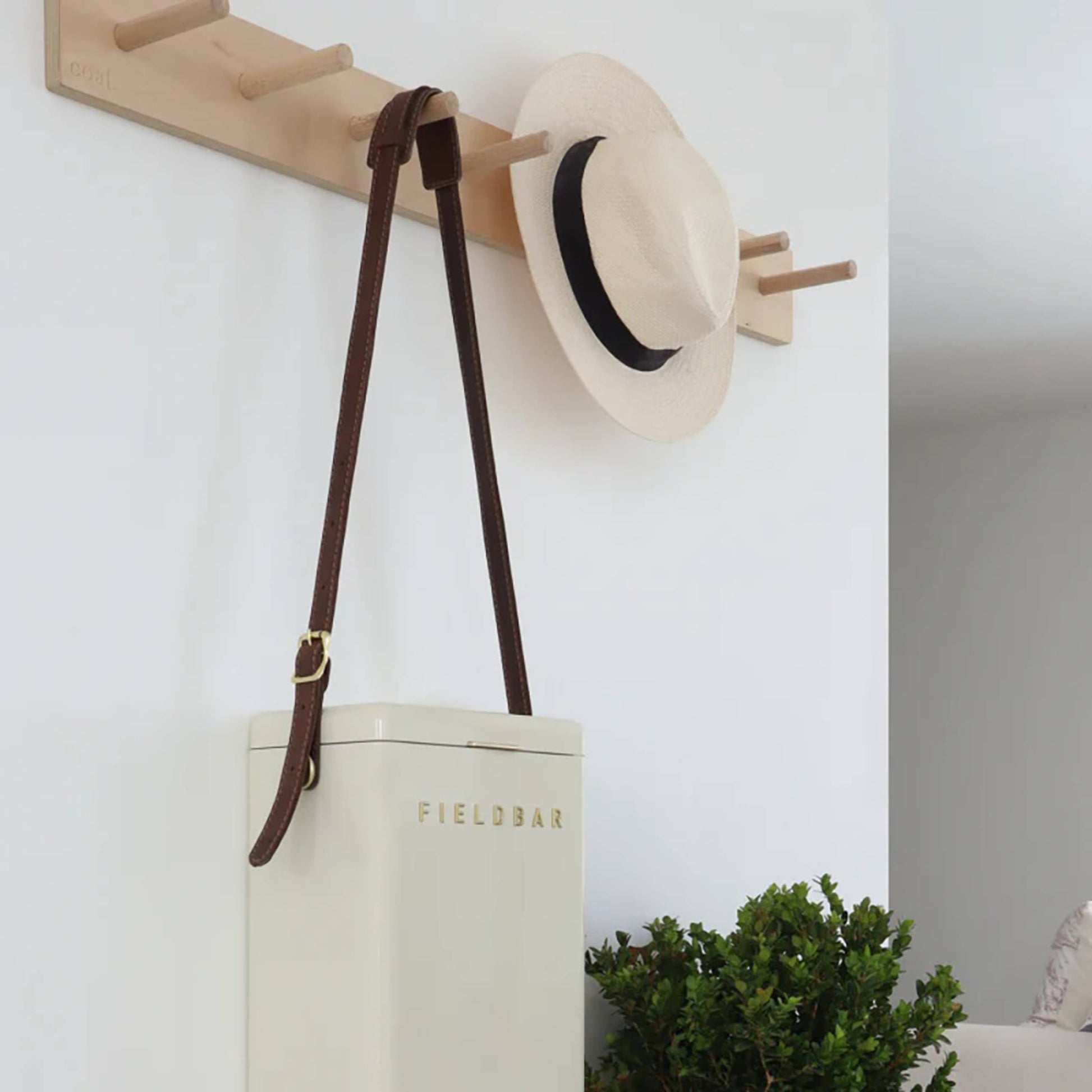 A beige Fieldbar Drinks Box Sling by Fieldbar with a premium leather strap hangs hands-free from a wooden wall peg rack. Above it is a light straw hat with a black band, and below sits a leafy green plant.