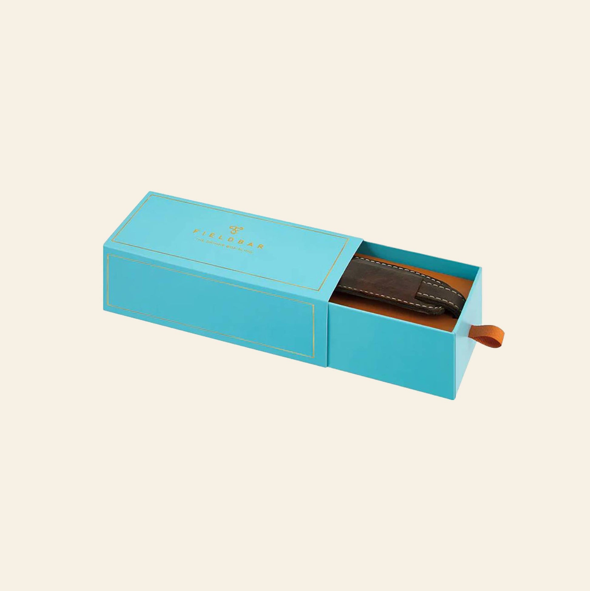 A turquoise rectangular Fieldbar box with gold lettering is partially open, revealing a premium leather Fieldbar Drinks Box Sling inside, featuring visible stitching and a pull tab for a stylish hands-free carrying solution.