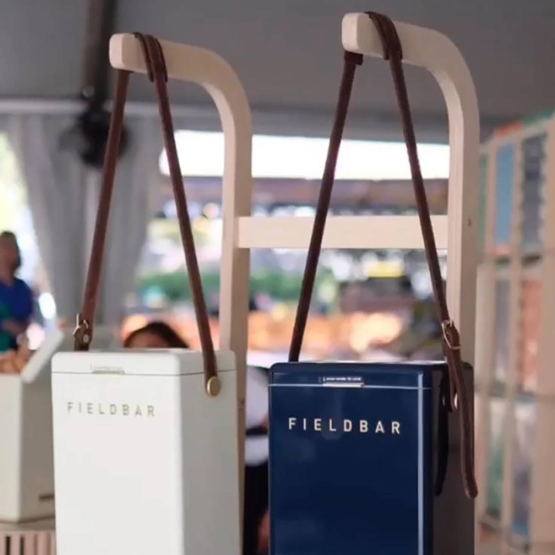 Two Fieldbar Drinks Box Slings—one white, one navy blue—hang by premium leather straps on a light wooden rack in a bright indoor space, offering a stylish hands-free solution for any gathering.