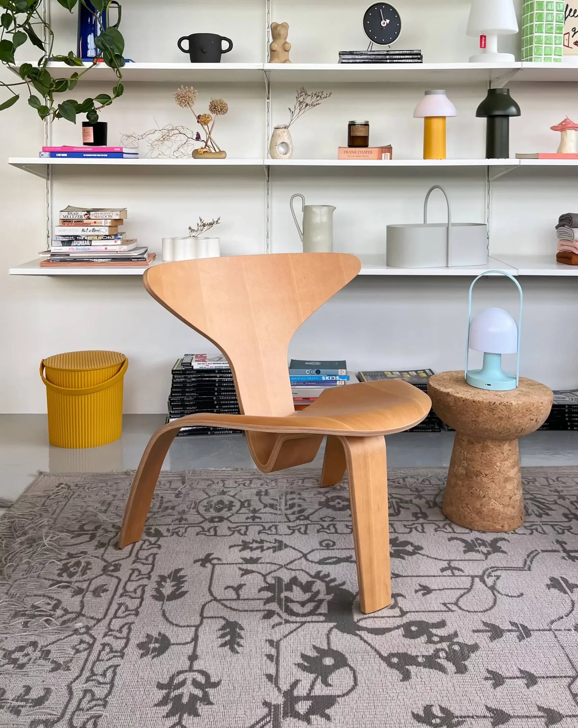 A Fritz Hansen PK0 A Lounge Chair sits on a patterned rug in front of white shelves filled with books, vases, décor, and plants. Nearby are a cork side table with a lamp and a yellow trash bin to the left.