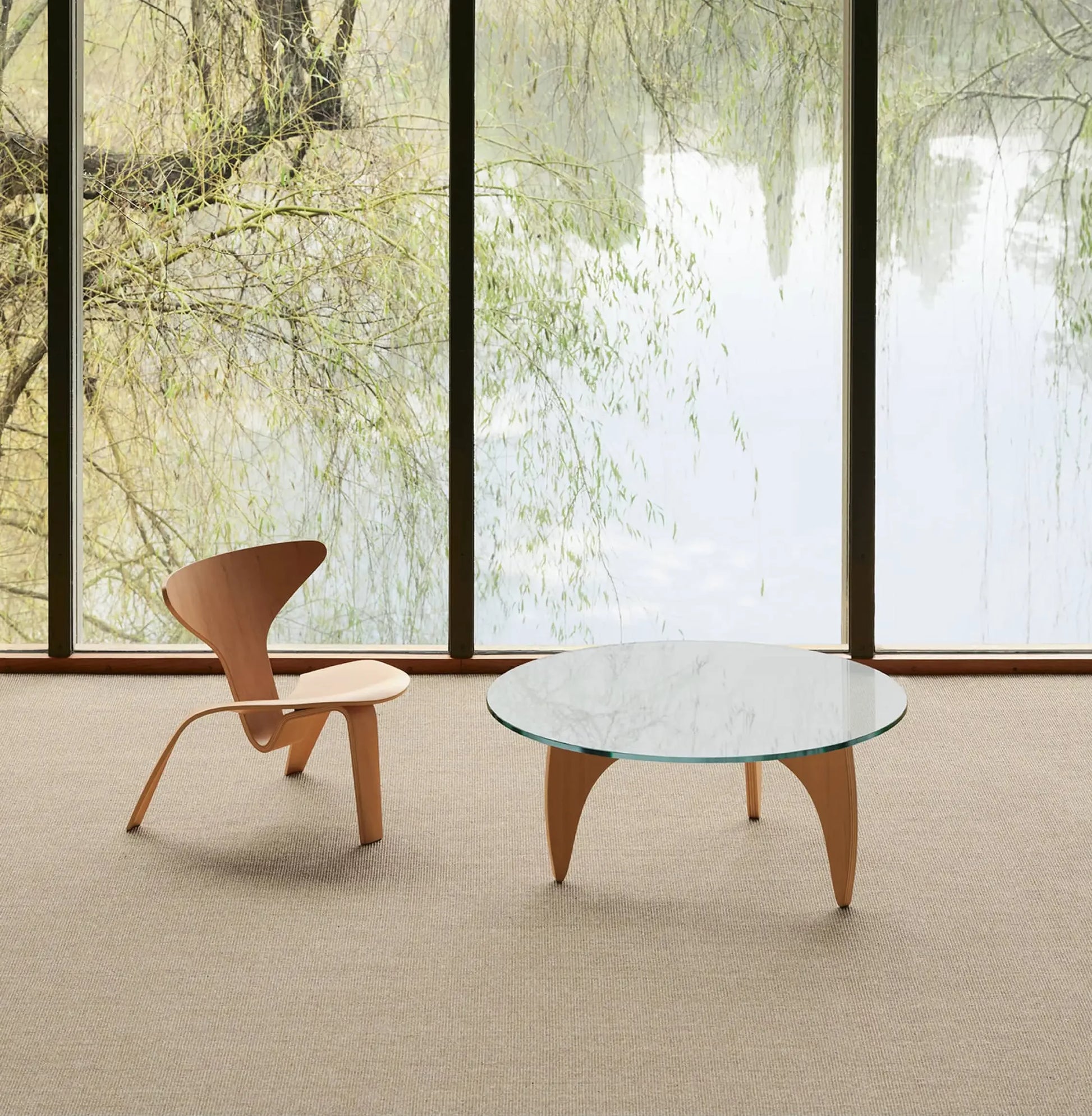 A minimalist room with a light wooden Fritz Hansen PK0 A Lounge Chair and a round glass coffee table on a beige carpet, set before floor-to-ceiling windows overlooking a serene, tree-lined lake—a modern design showcase.
